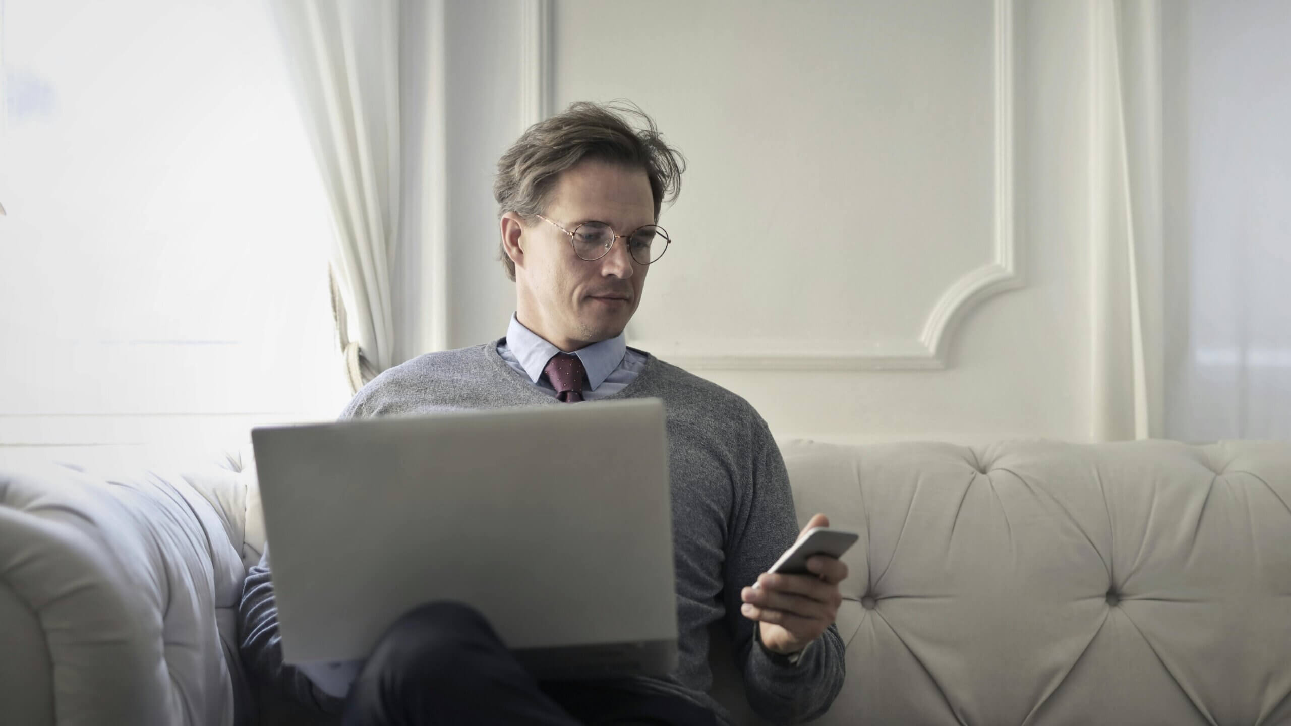 Man in glasses and tie using laptop and smartphone while sitting on a white couch in a bright room