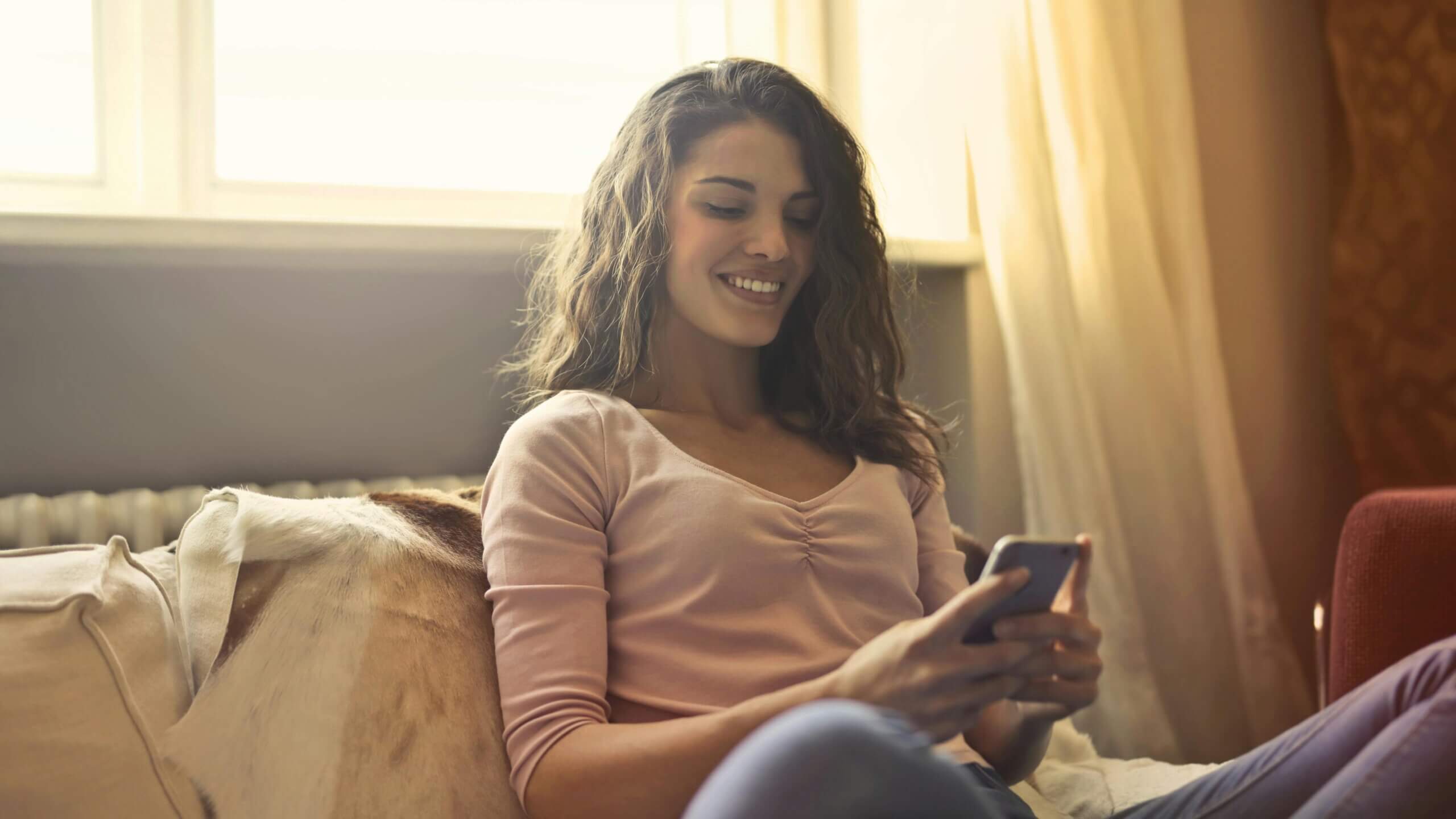 Young woman smiling while using a smartphone on a couch near a window with natural light