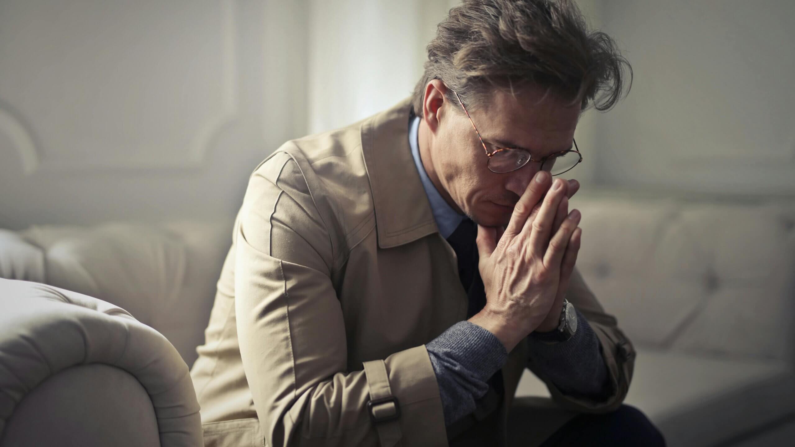 Man in glasses and beige coat sitting on a couch with hands pressed together, appearing deep in thought or prayer.