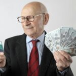 Elderly man in suit holding a credit card in one hand and fanned US $100 bills in the other.