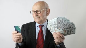 Elderly man in suit holding a credit card in one hand and fanned US $100 bills in the other.