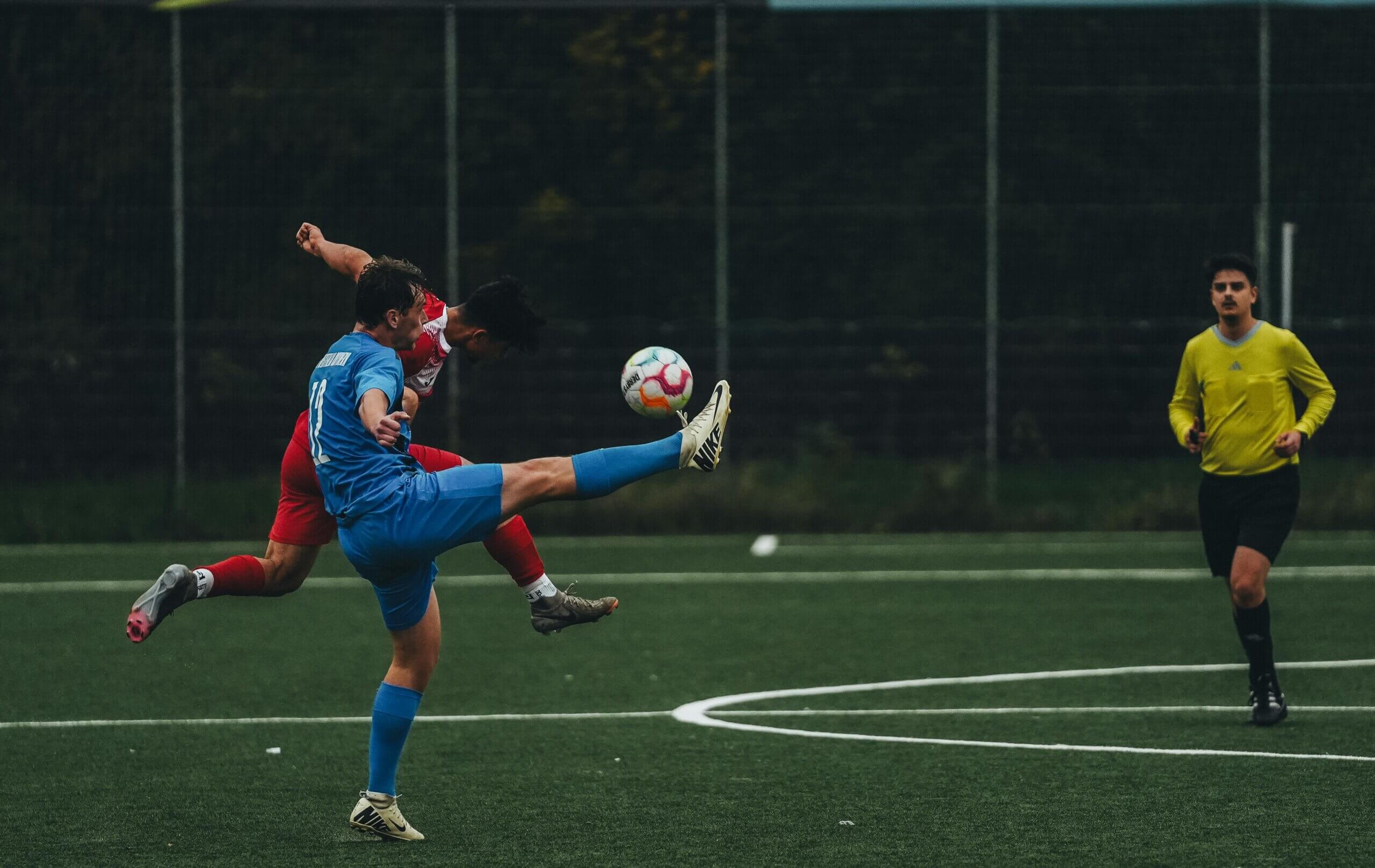 Two soccer players in blue and red jerseys competing for the ball with a referee in yellow nearby on the field.