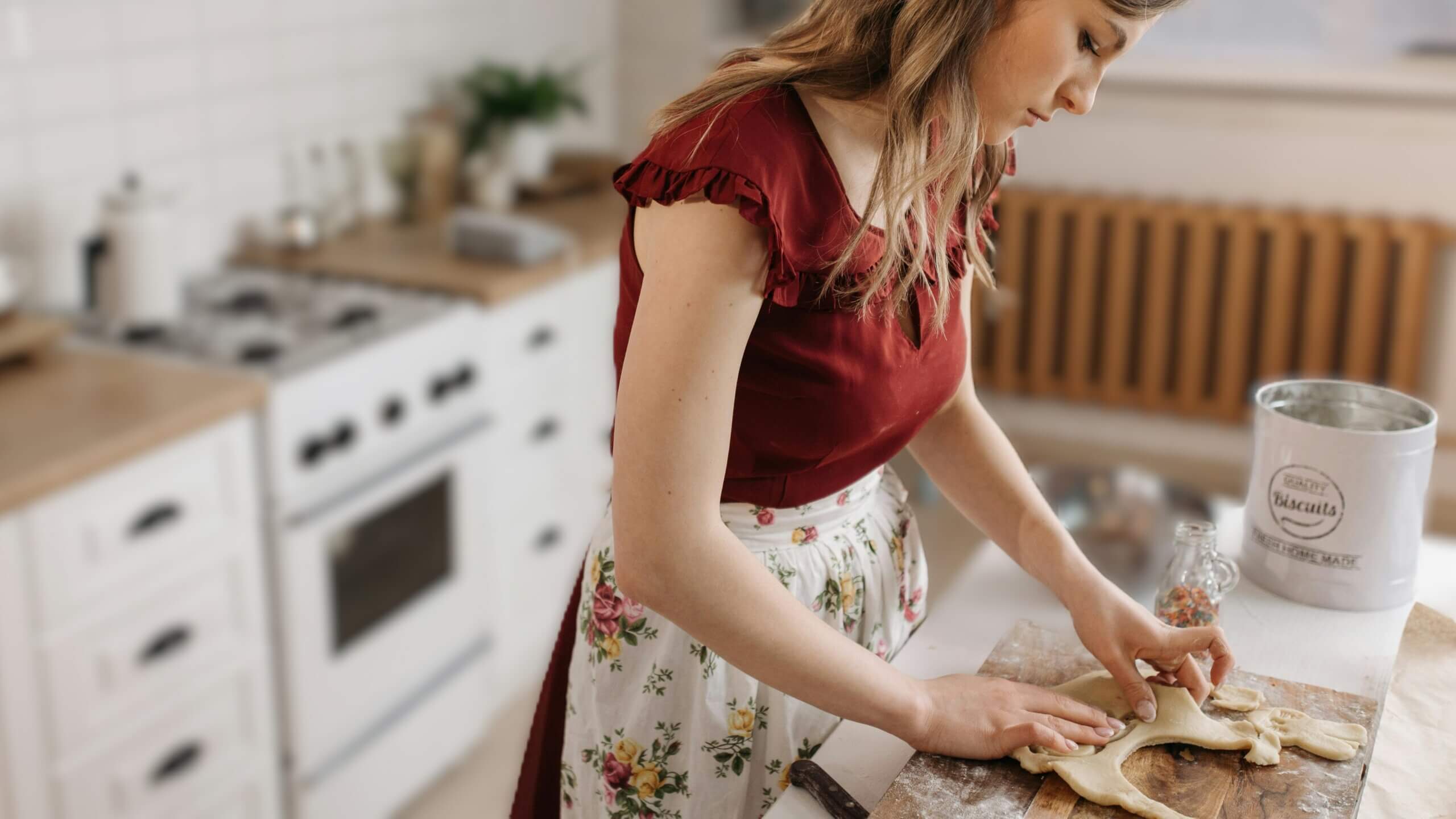 Woman in red top and floral apron shaping dough on a wooden board in a kitchen