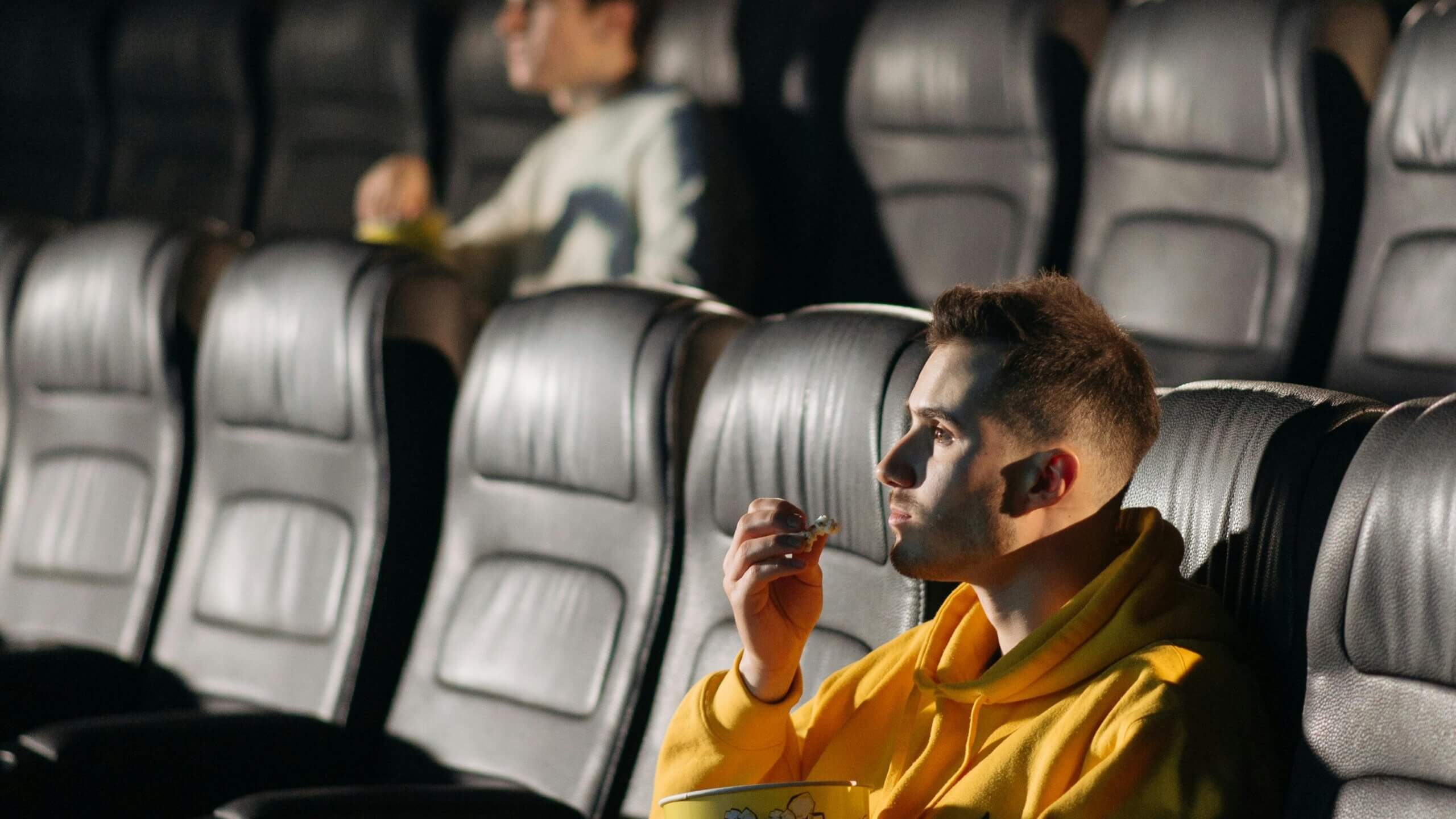 Young man in a yellow hoodie eating popcorn while watching a movie in a nearly empty theater.