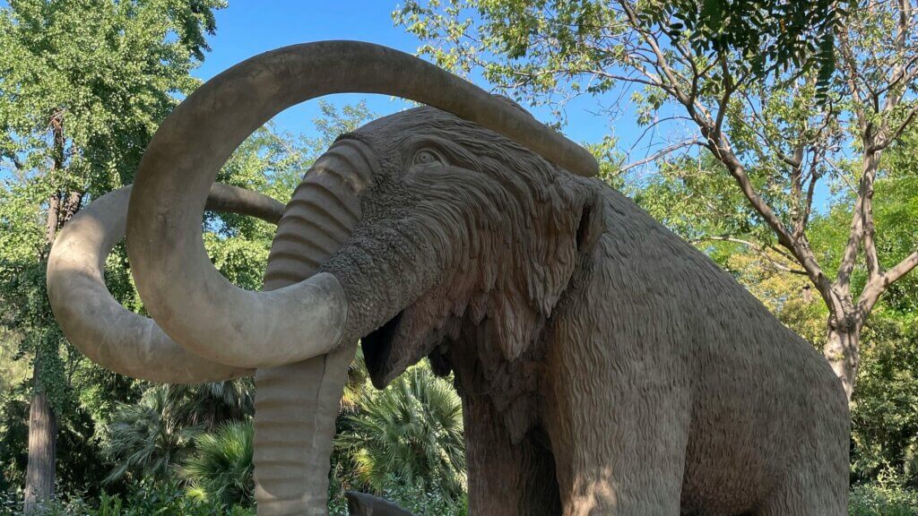 Large woolly mammoth statue with curved tusks surrounded by trees under a clear blue sky