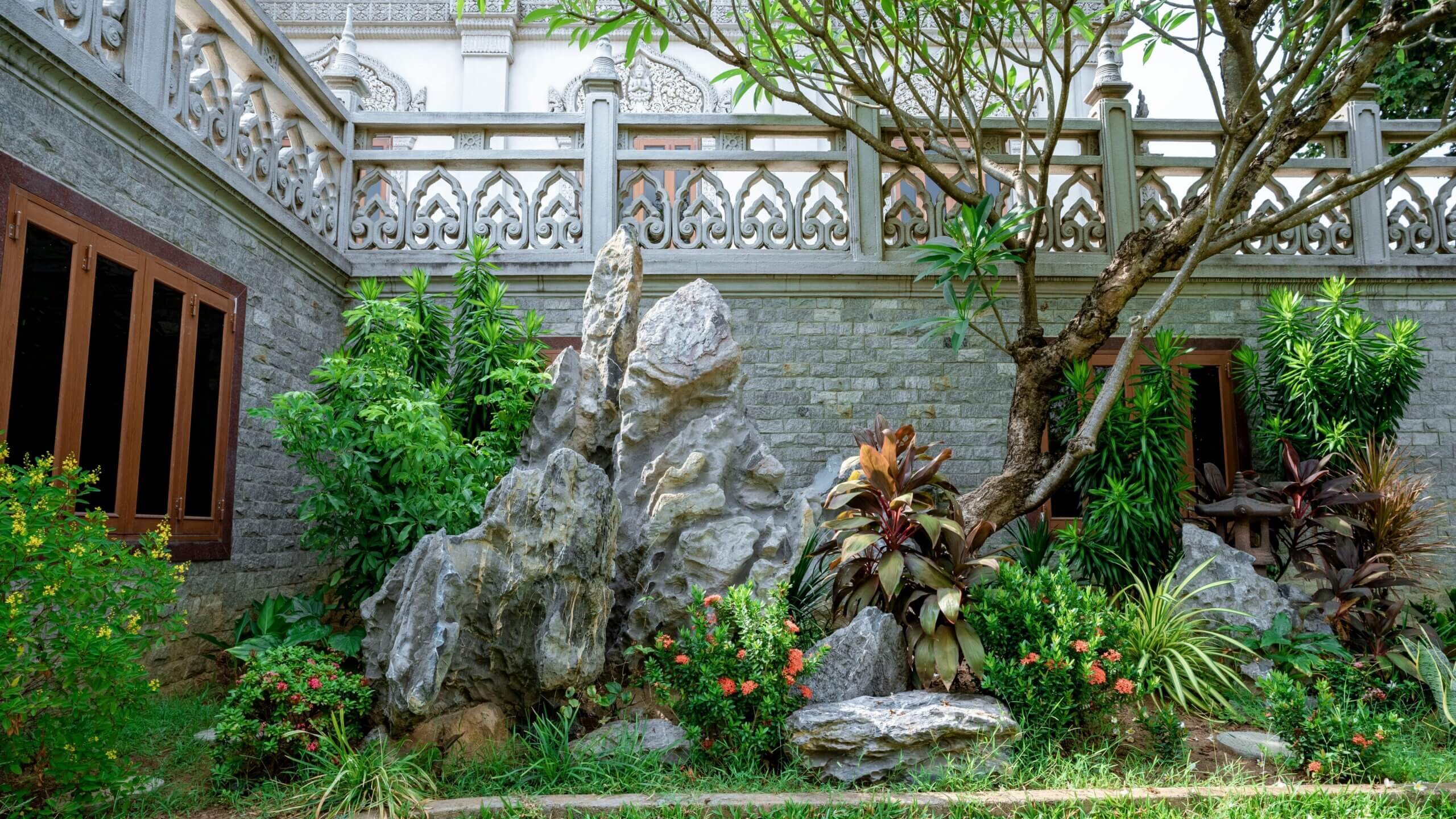 Rock garden with various plants and a tree in front of a stone wall with decorative railing and wooden windows