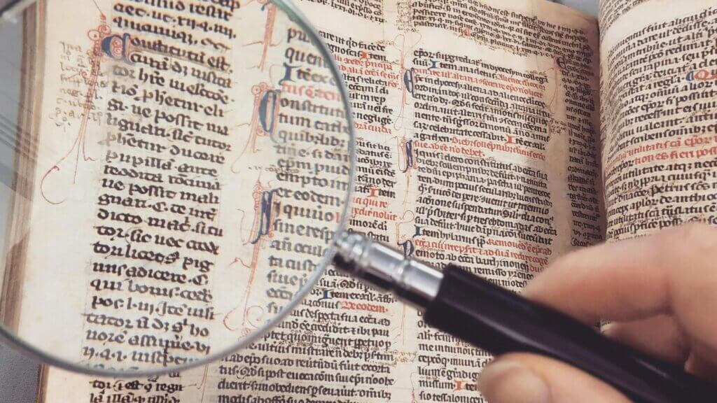 Hand holding magnifying glass over an ancient manuscript with black and red Gothic script text.