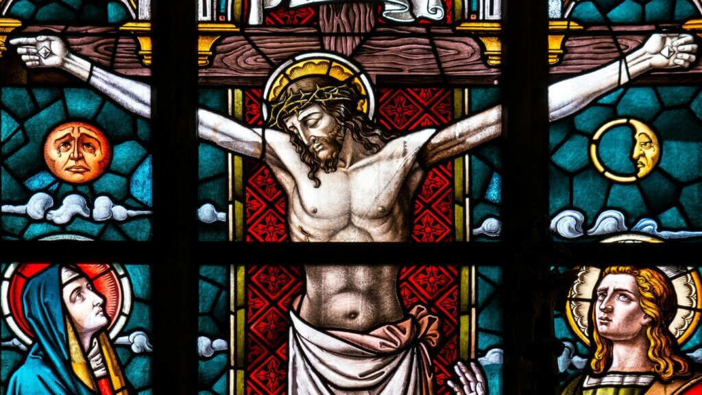 Stained glass depicting Jesus Christ crucified with Mary and an angel below, with sun and moon symbols above.