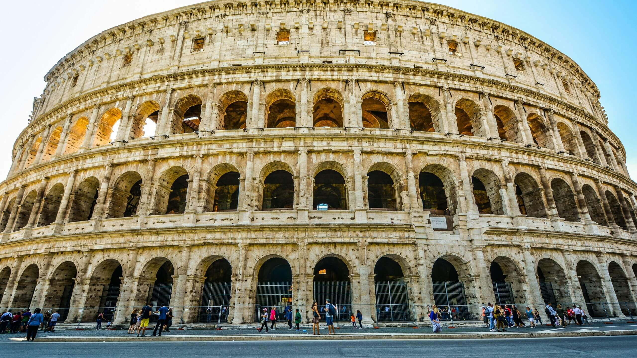 The Colosseum in Rome with tourists walking and standing near its arches on a clear day.