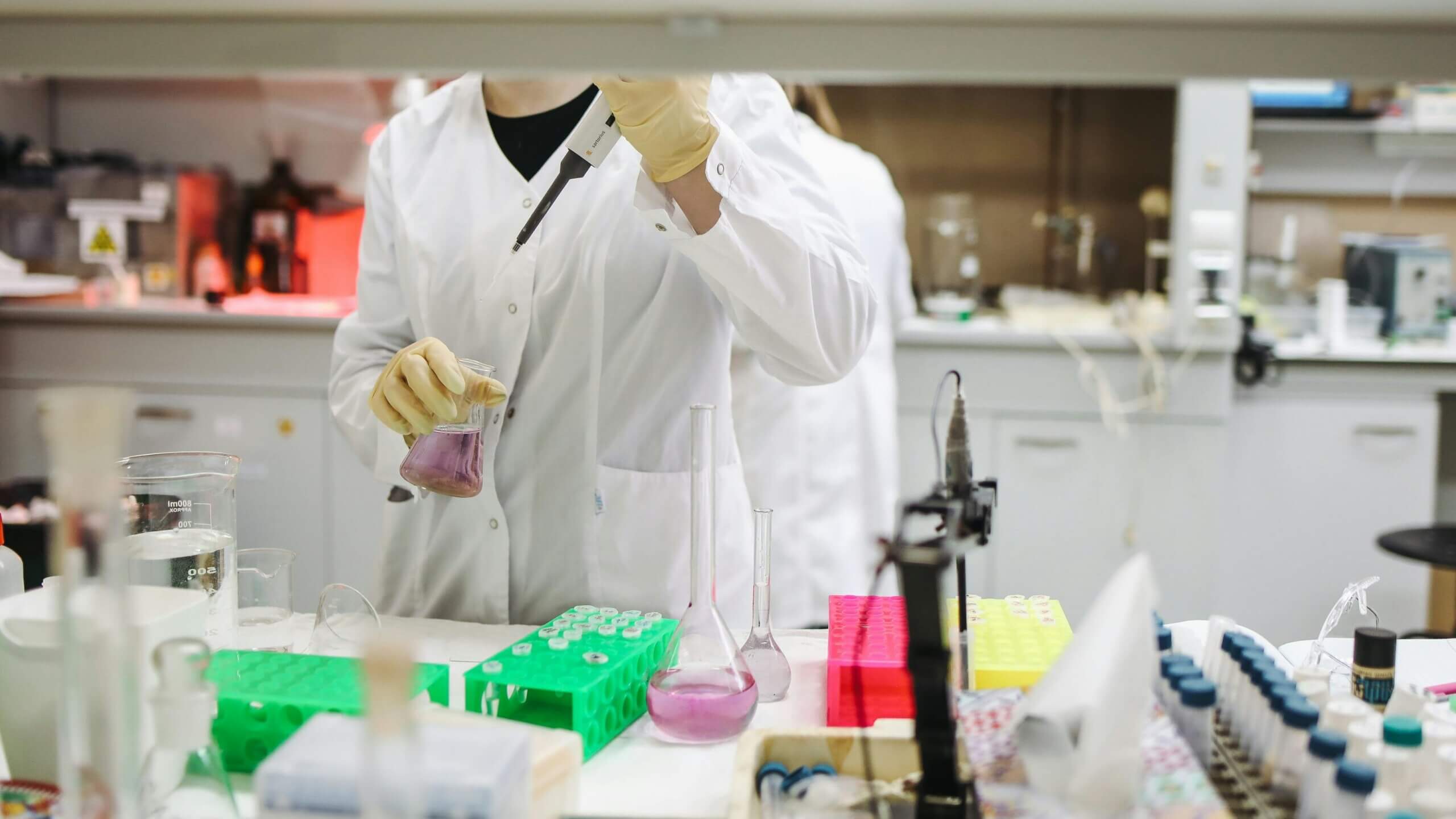 Scientist in white lab coat using a pipette to transfer purple liquid into a flask in a laboratory.