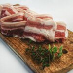 Raw pork belly slices on a wooden cutting board with fresh thyme sprigs on a white surface