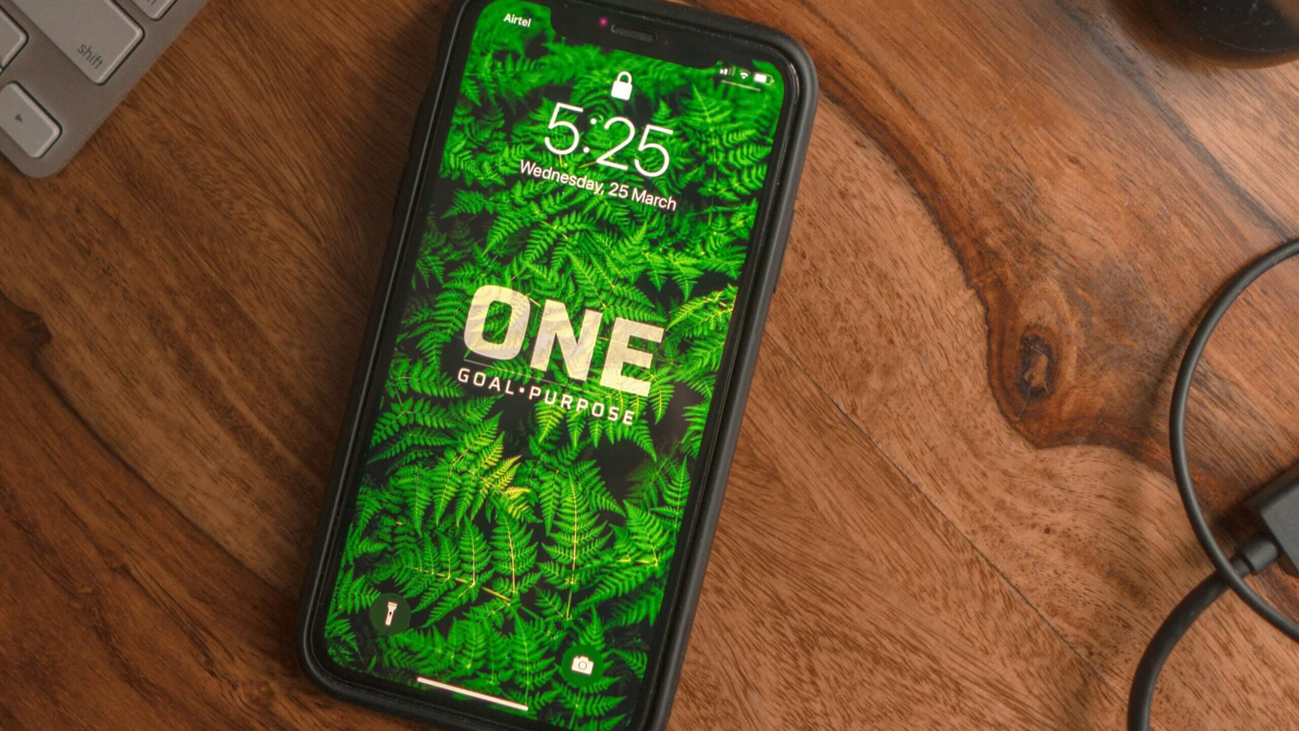 Smartphone on wooden table displaying lock screen with green fern wallpaper and text "ONE GOAL PURPOSE"