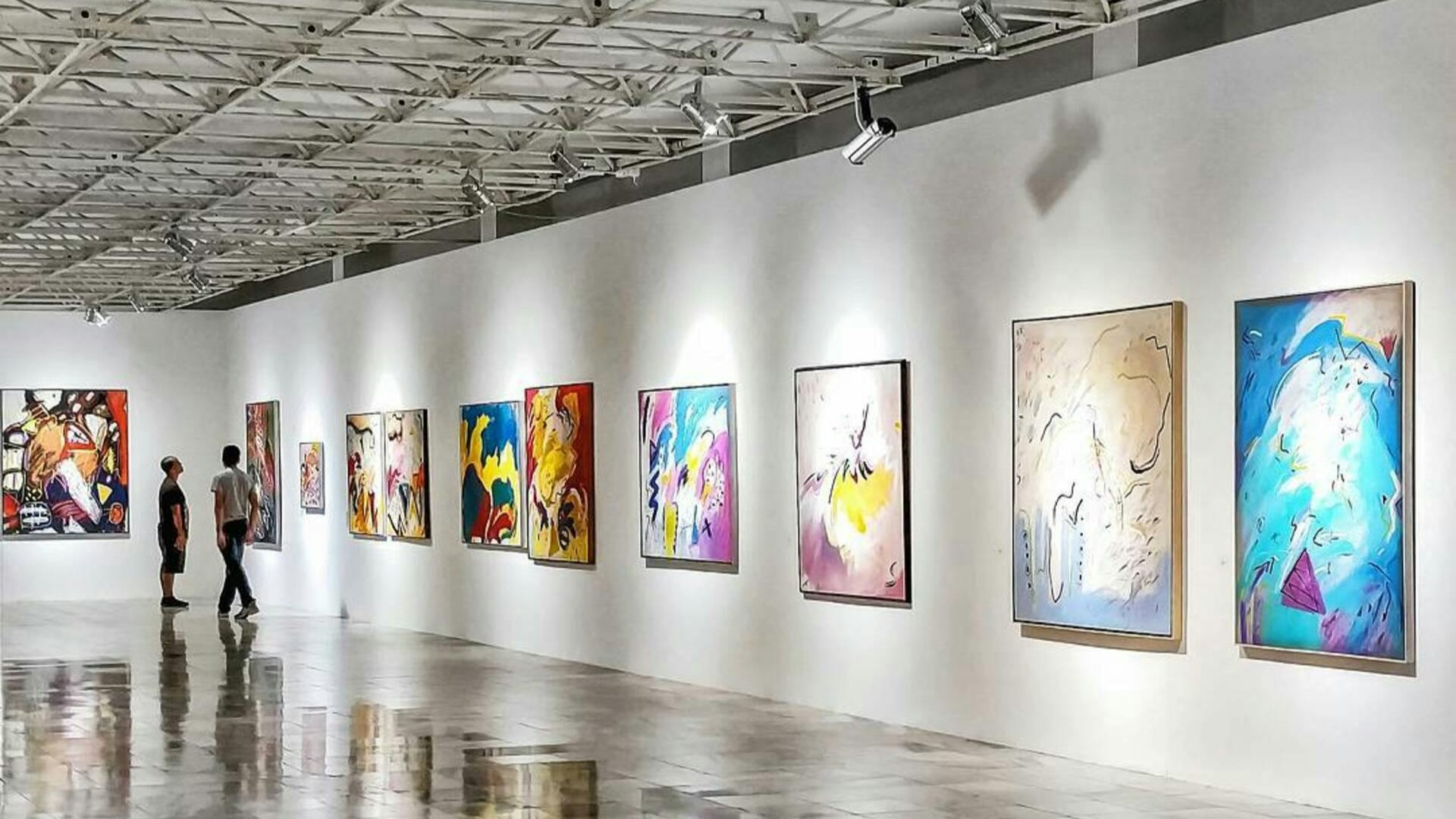 Two people viewing colorful abstract paintings in a modern art gallery with white walls and polished floors