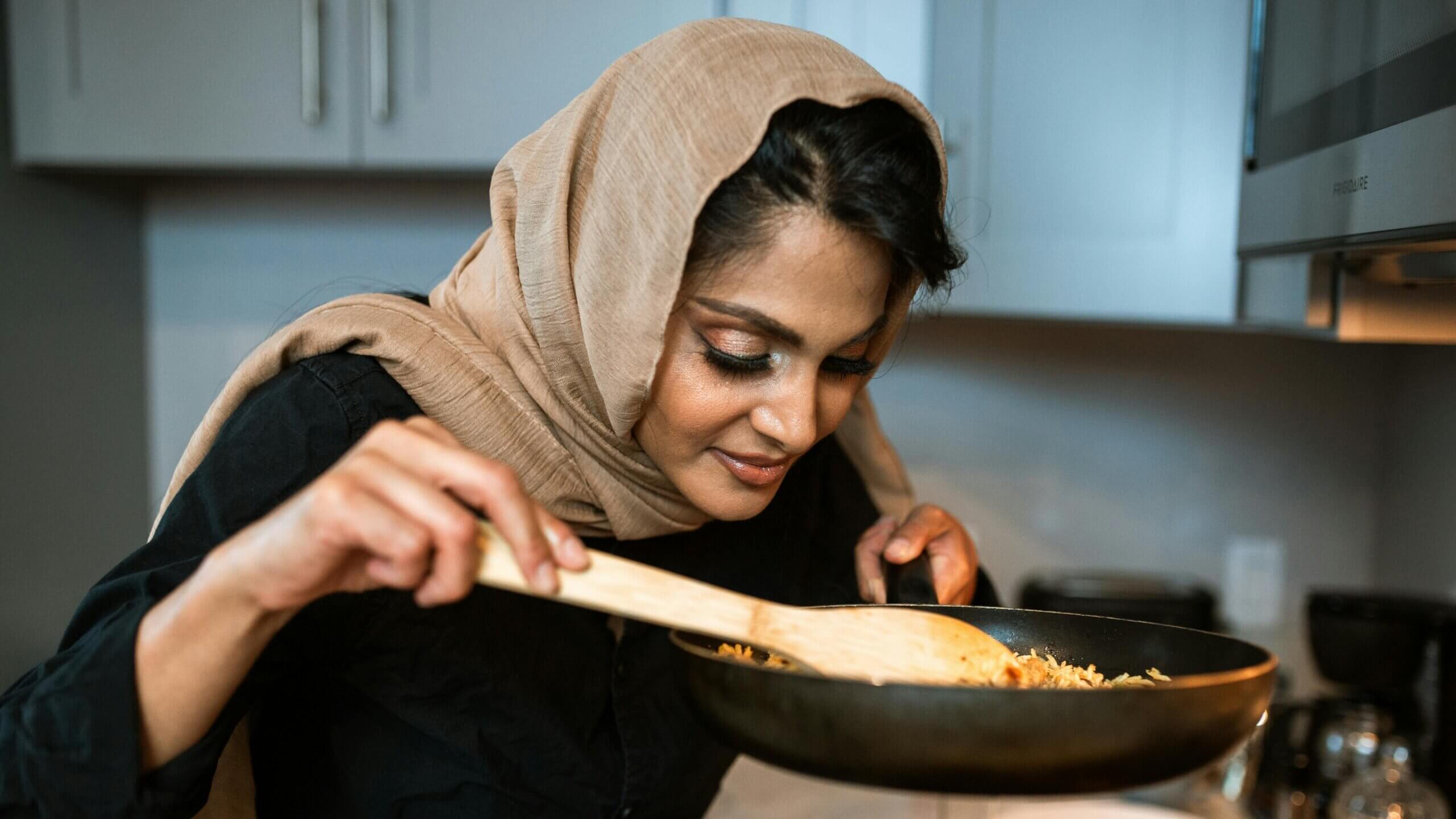 Woman wearing a beige hijab smelling food in a frying pan while cooking in a kitchen