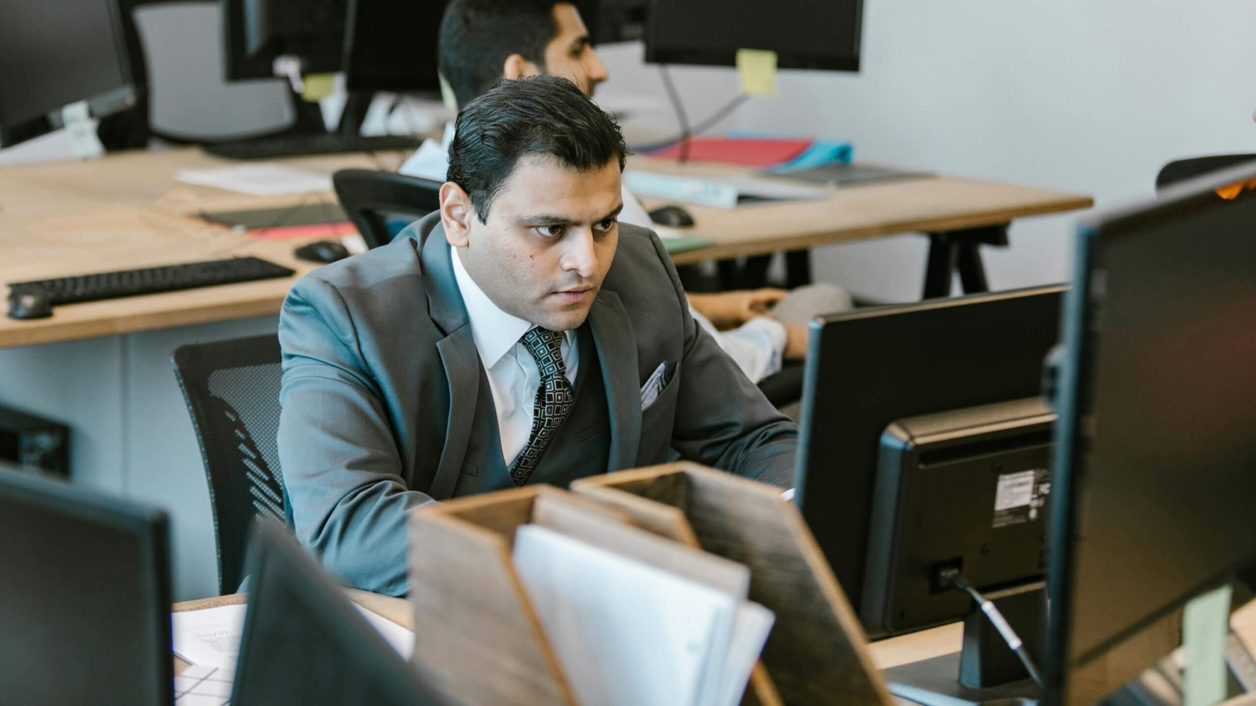 Man in a gray suit and patterned tie focused on a computer screen in an office setting.