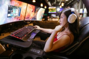 Woman wearing white headphones gaming on a triple-monitor setup with a red backlit keyboard in a dark room