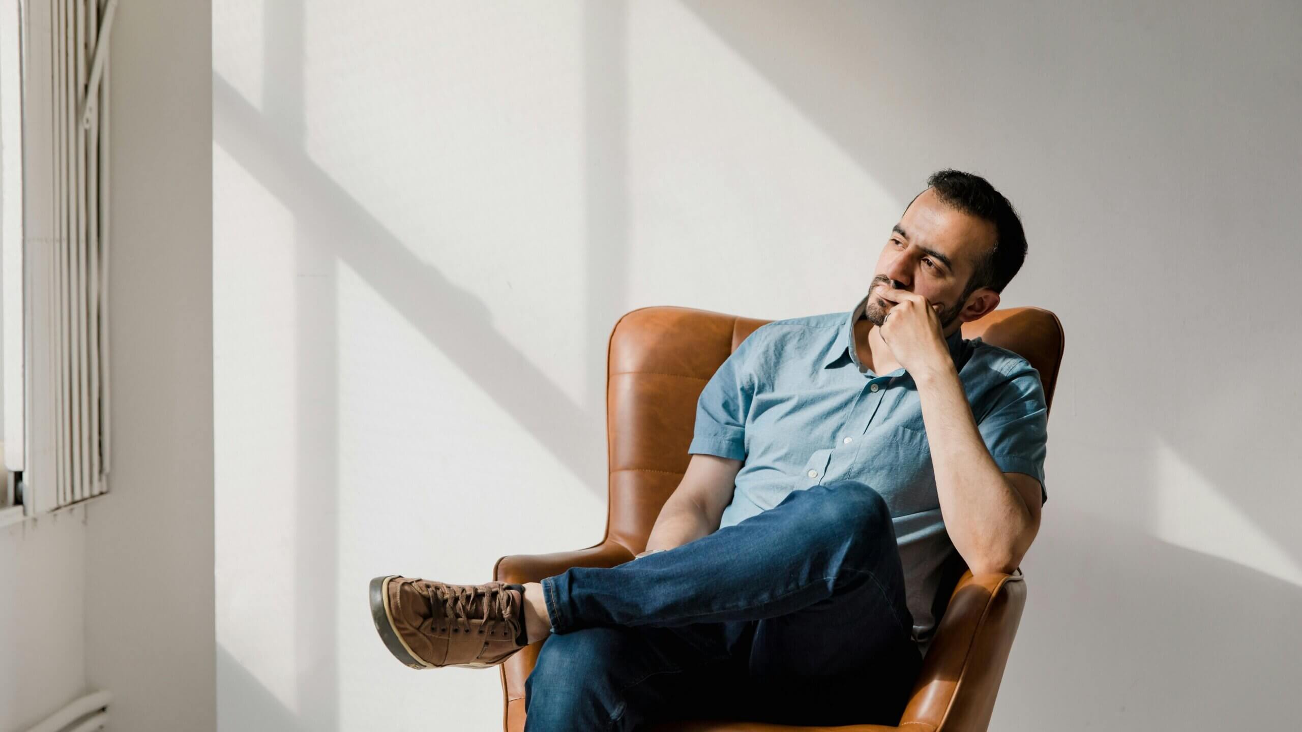 Man in blue shirt and jeans sitting thoughtfully in a brown leather chair near a window with sunlight patterns on the wall