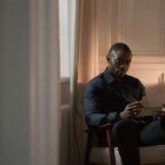Man in dark shirt sitting in chair reading a letter in a softly lit room with books and a lamp on a side table