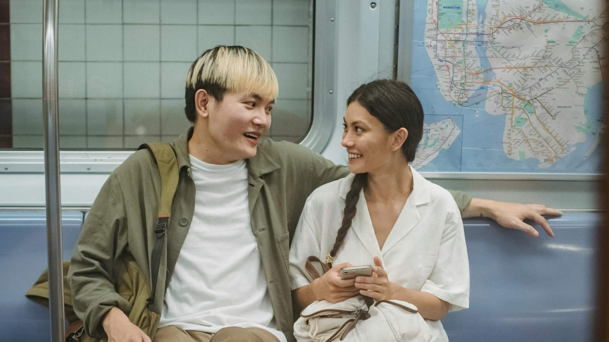 Two people smiling and talking while sitting on a subway train with a map of Manhattan in the background.