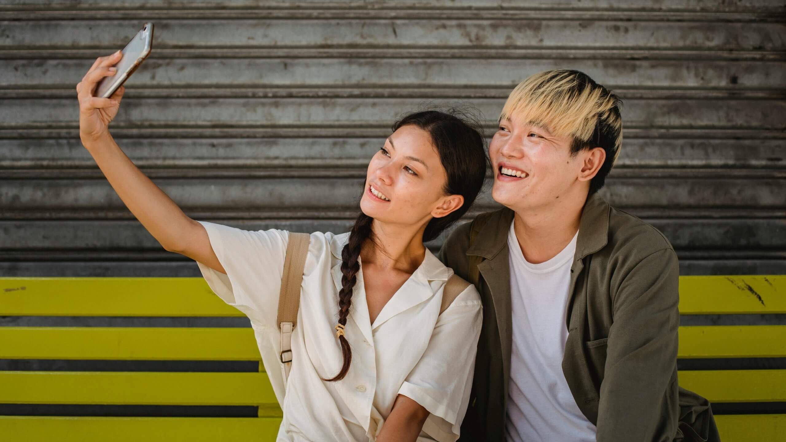 Two young people sitting on a yellow bench smiling and taking a selfie with a smartphone.