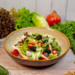 Mixed green salad with strawberries, blackberries, blueberries, and almonds in a bowl surrounded by fresh vegetables and fruits
