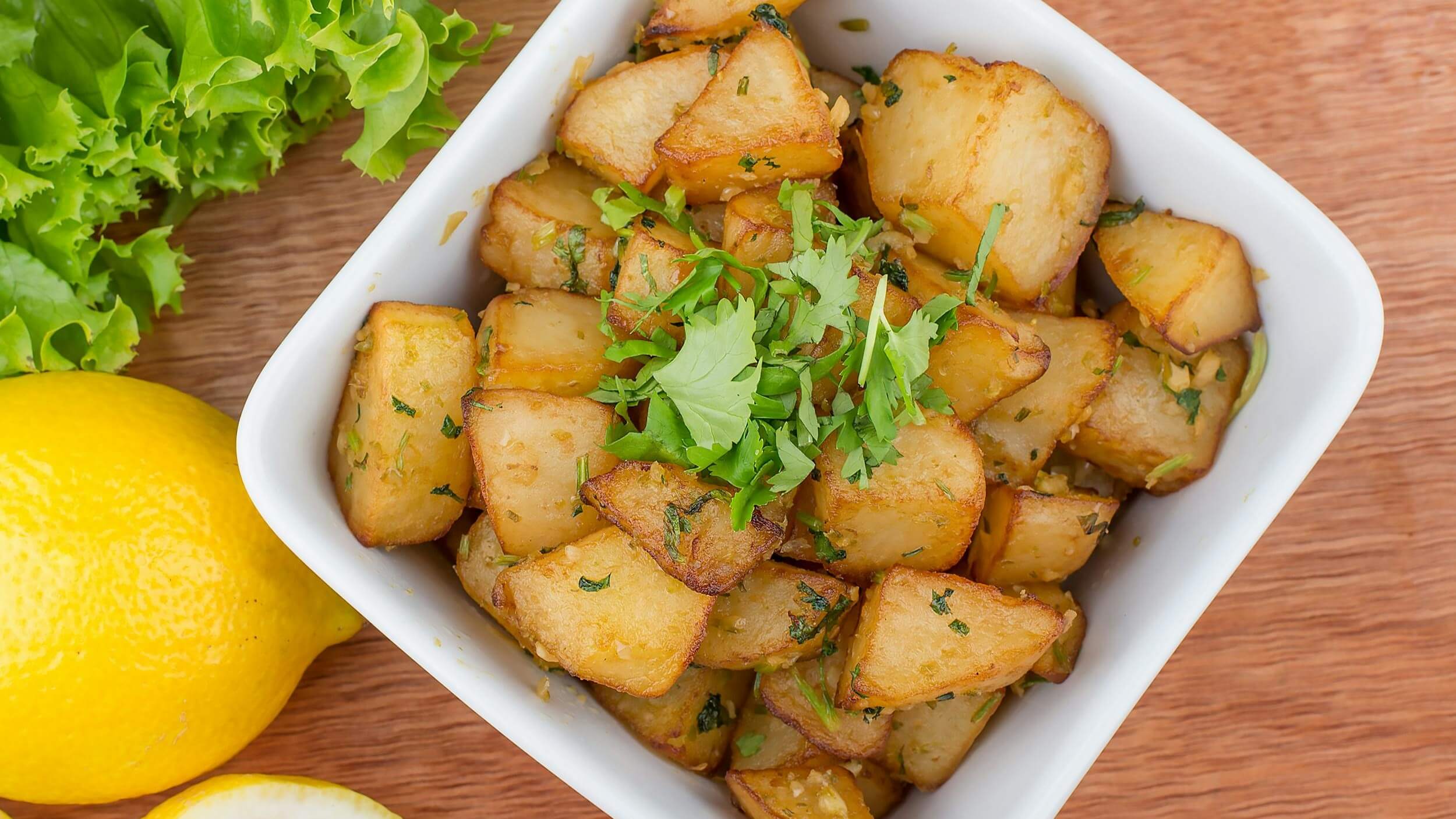 Roasted potato chunks garnished with fresh cilantro in a white bowl with lemon and lettuce on the side