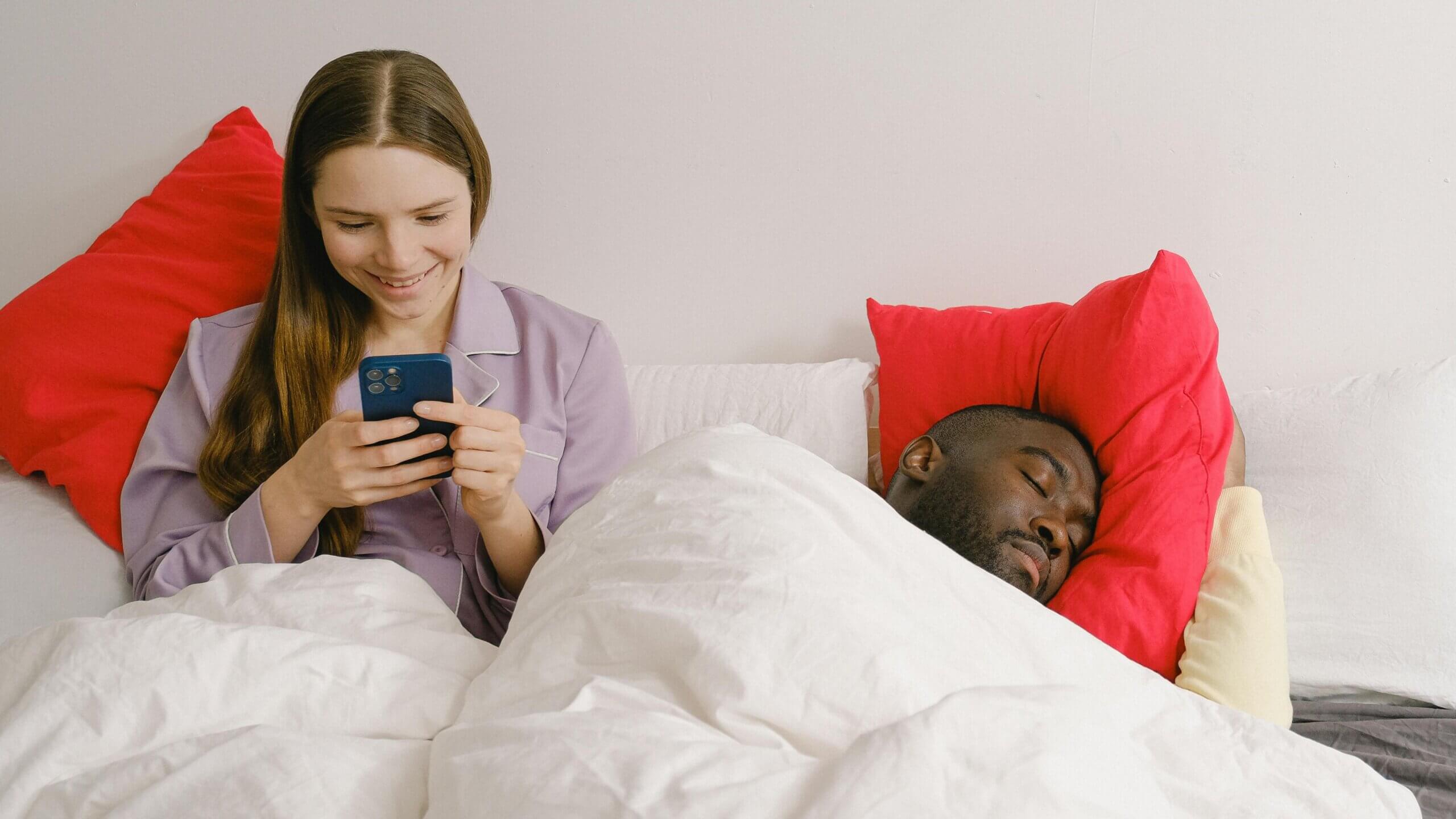 Woman in purple pajamas using a smartphone in bed while a man sleeps beside her on a red pillow