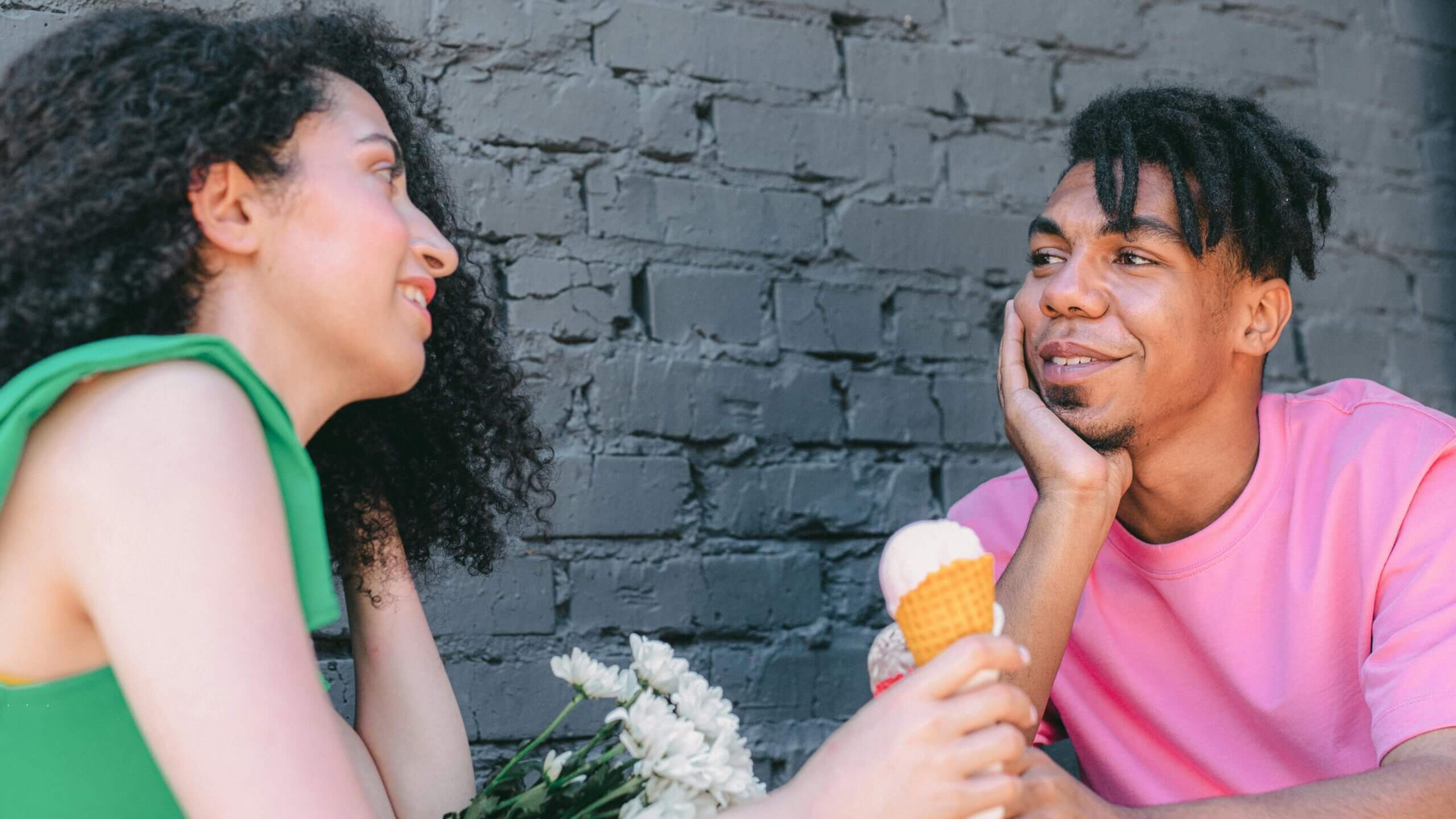 Young couple smiling and holding ice cream cones while sitting against a gray brick wall