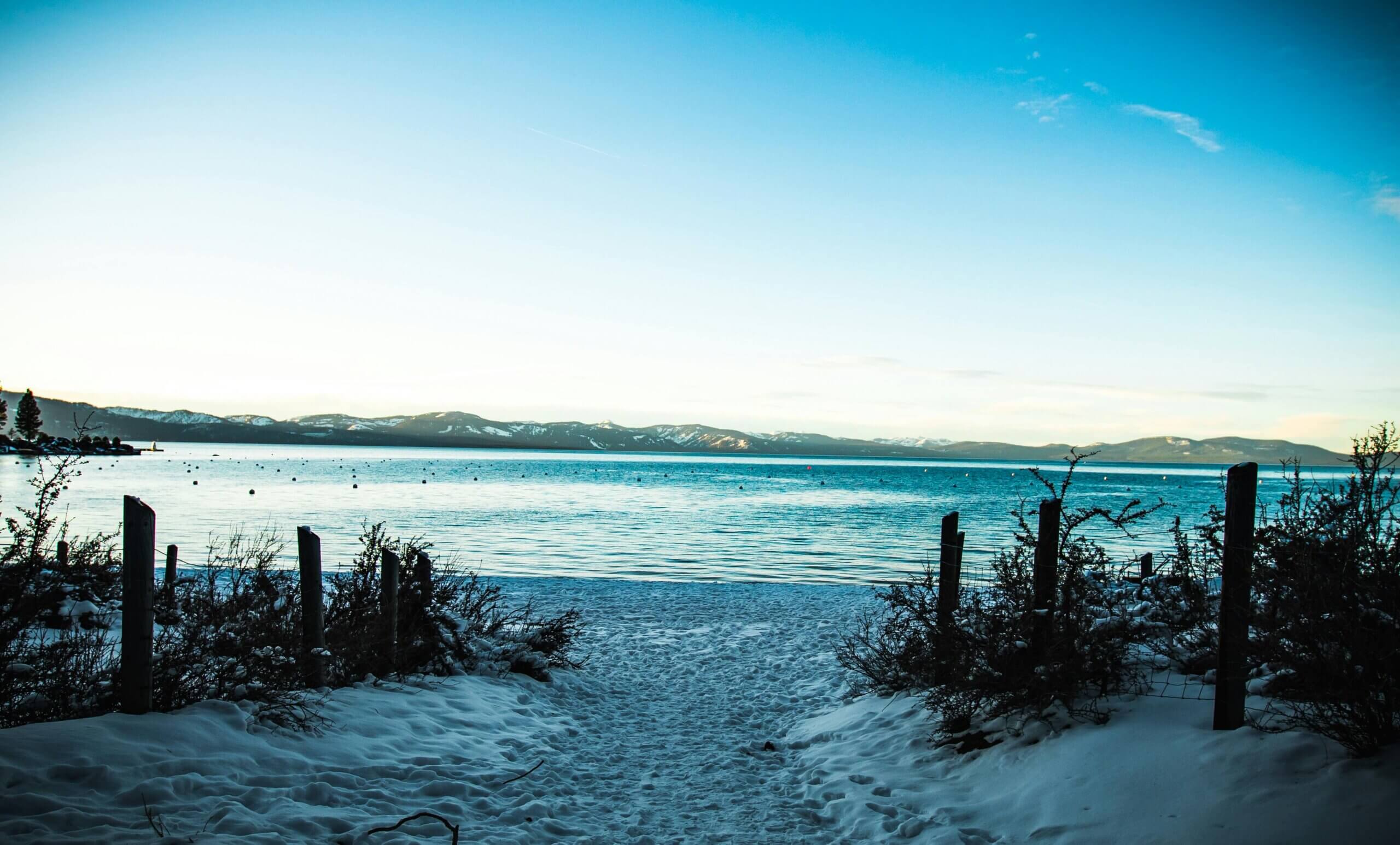 Snow-covered path leading to a lake with mountains in the background under a clear blue sky