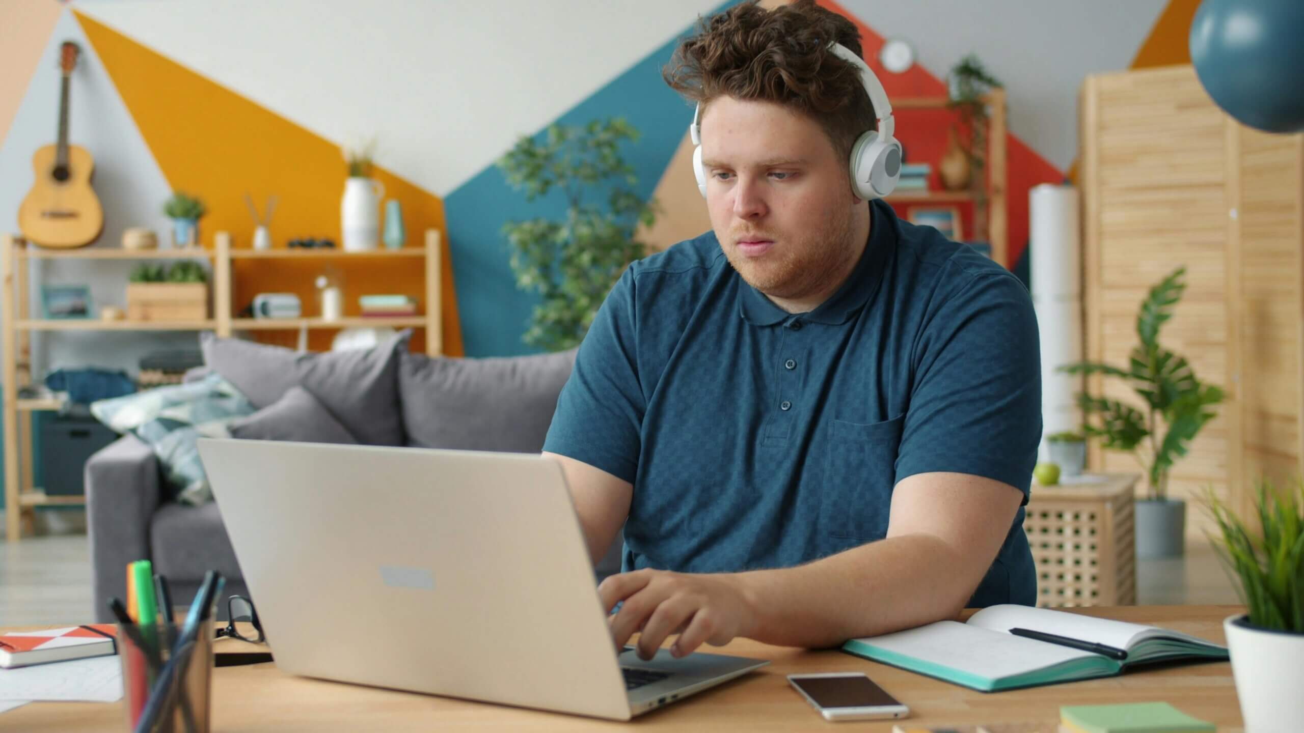Man wearing white headphones working on a laptop at a desk with a notebook and smartphone in a colorful room