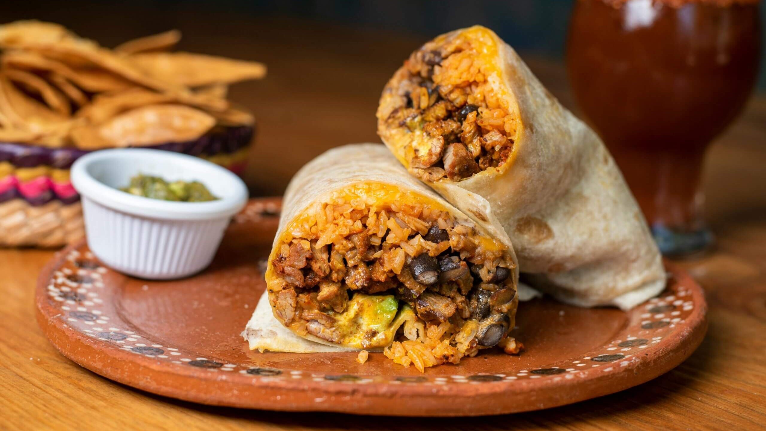 Two halves of a burrito filled with rice, black beans, meat, and cheese on a clay plate with chips, salsa, and a michelada drink.
