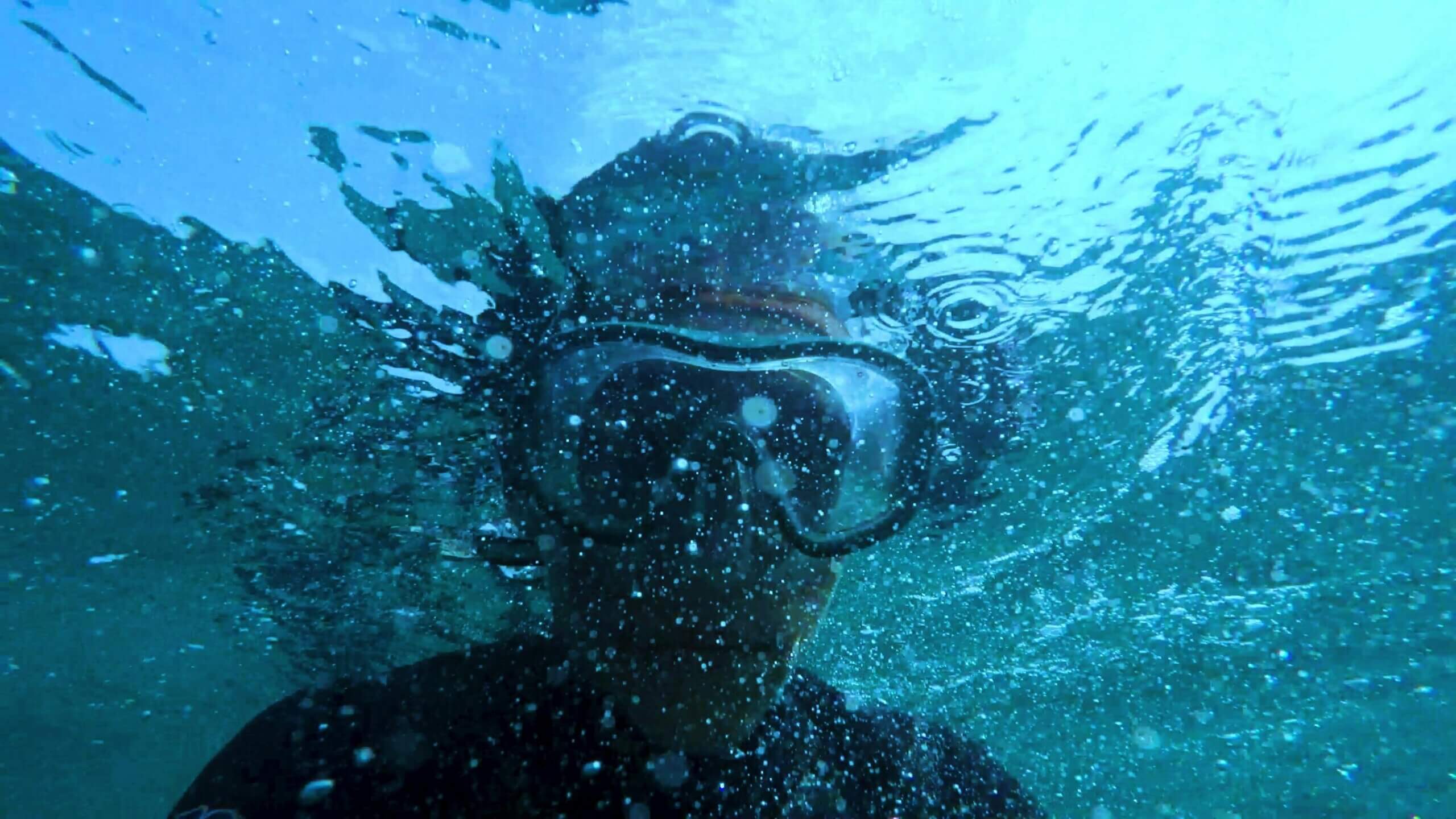 Person wearing a diving mask underwater surrounded by bubbles and ripples.