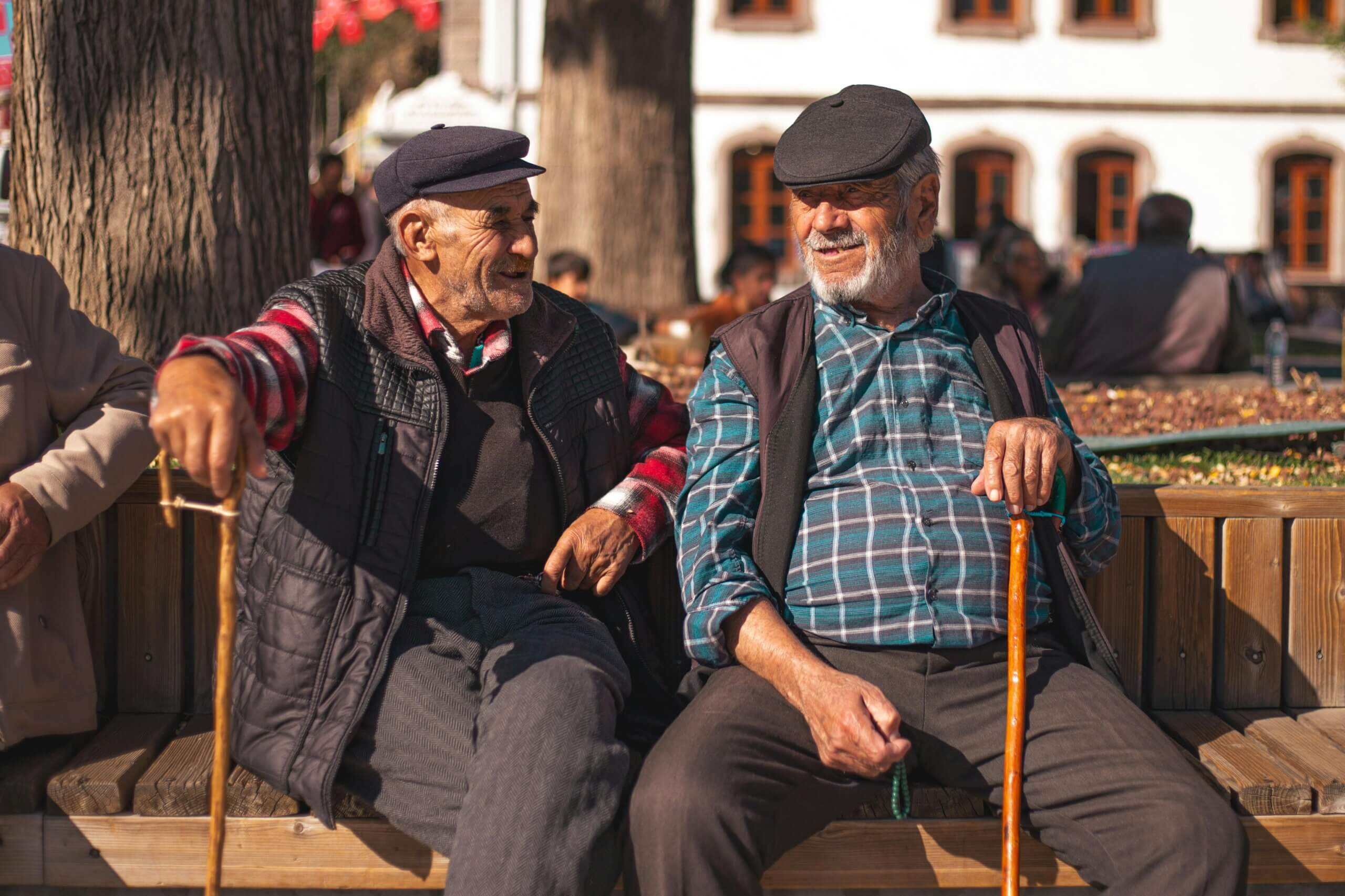 Two elderly men wearing flat caps and holding wooden canes sitting and chatting on a bench outdoors