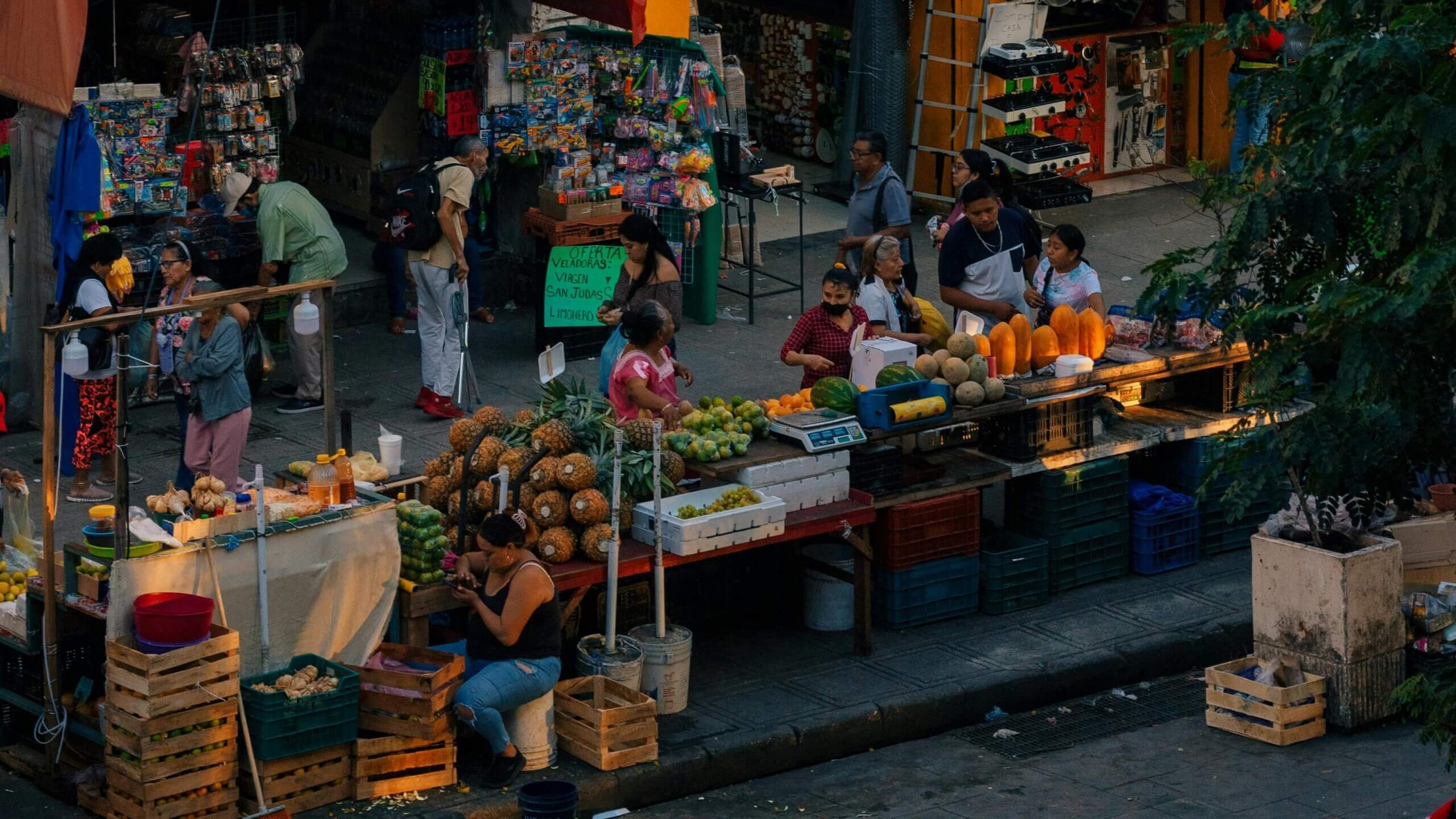 Street market scene with vendors selling pineapples, papayas, melons, and other fruits to customers.