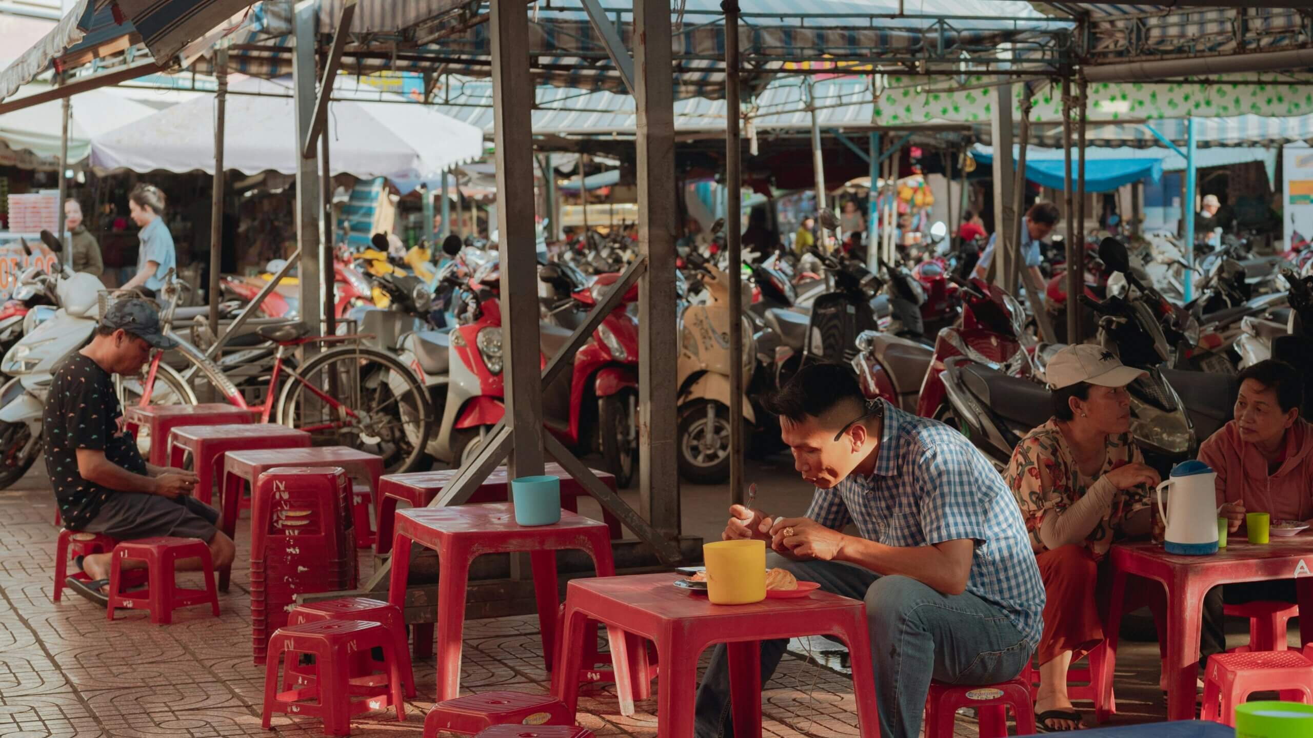 People sitting on small red stools eating and talking in an outdoor market with parked motorbikes in the background