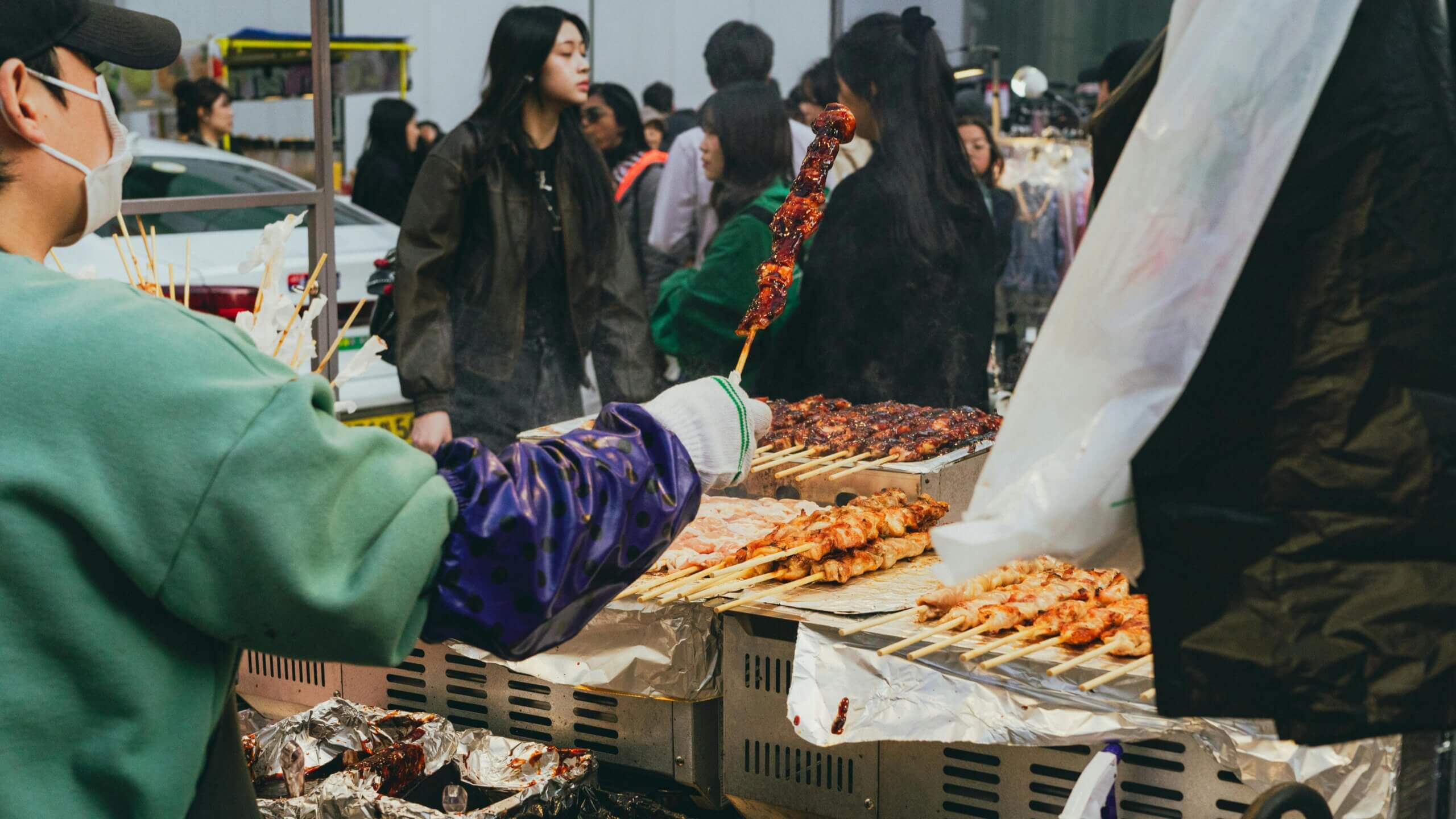 Street vendor grilling skewered meat at an outdoor market with customers in the background.