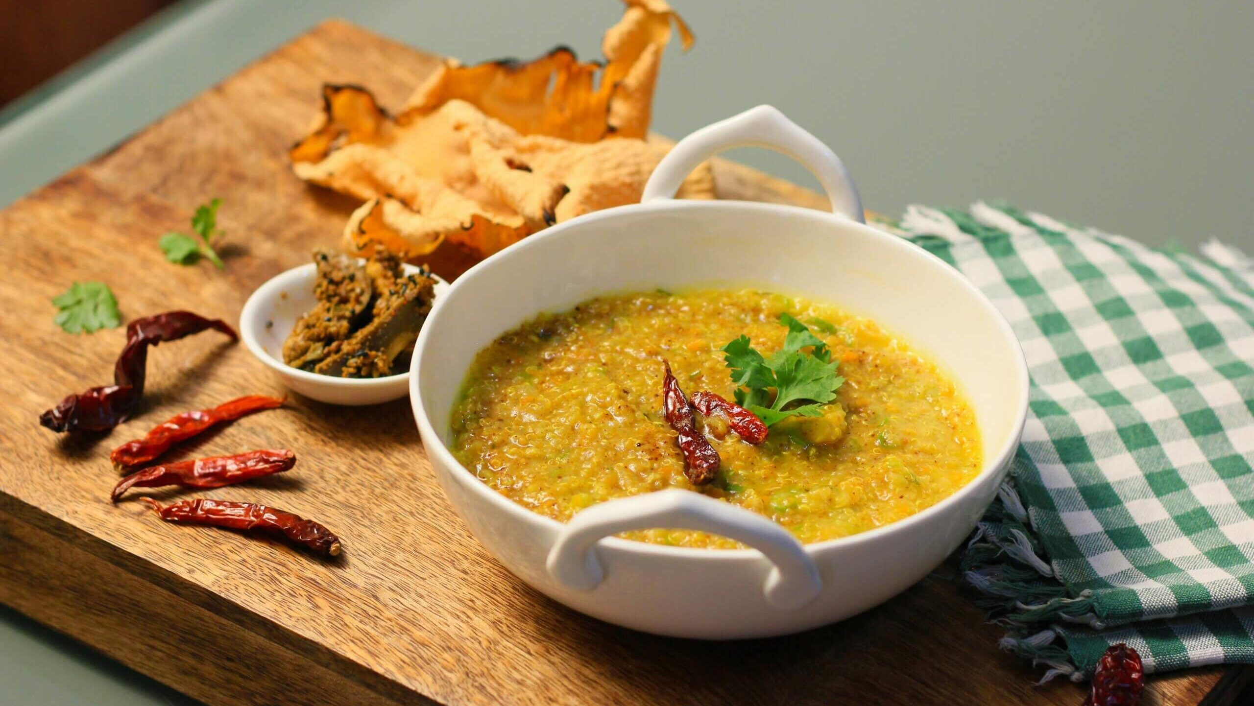 White bowl of Indian dal garnished with dried red chilies and cilantro on a wooden board with dried mushrooms and chutney.