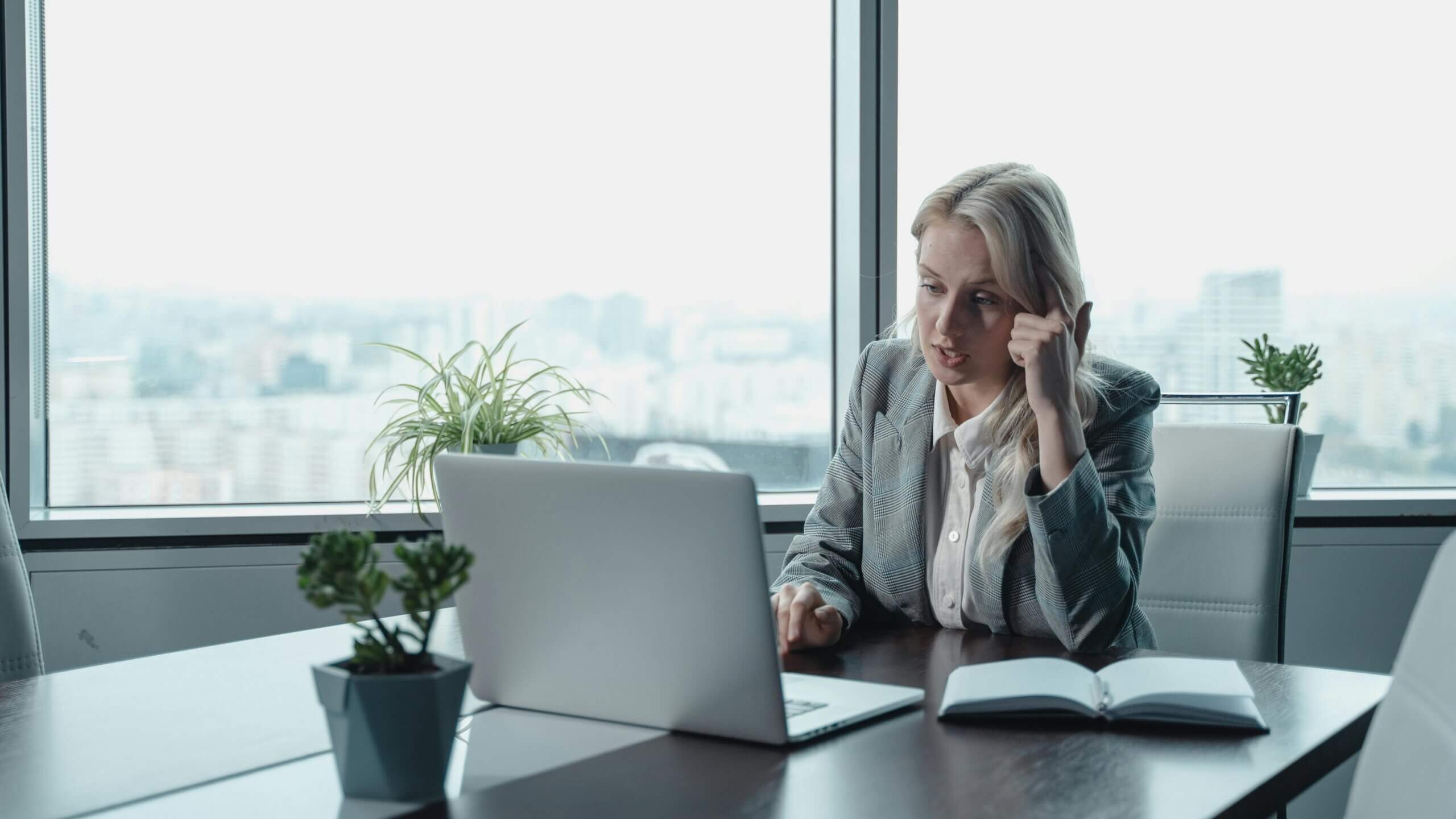 Blonde woman in a gray blazer working on a laptop at a conference table with plants and a notebook nearby