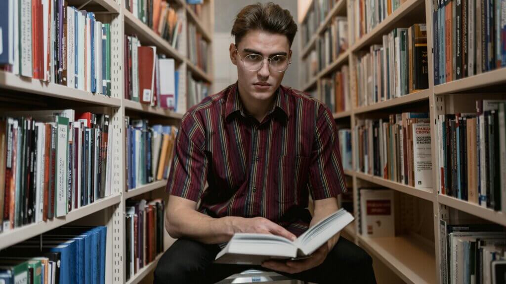 Young man wearing glasses and a striped shirt reading a book while sitting between library shelves