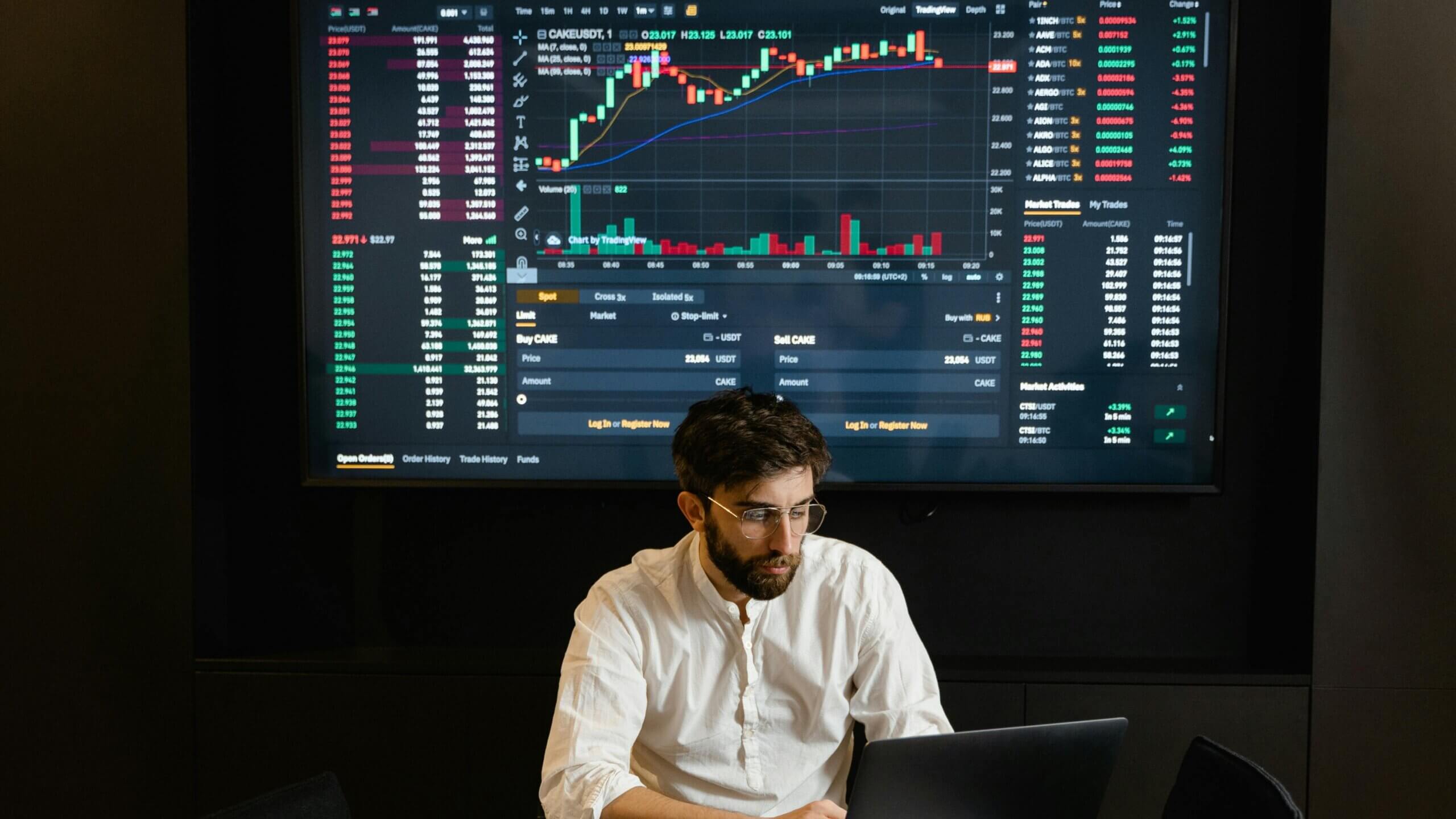 Man in white shirt using laptop in front of large screen displaying cryptocurrency trading charts and data
