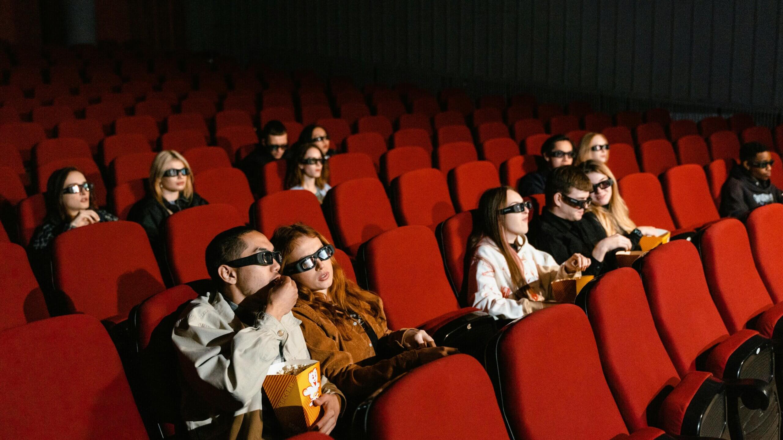 People wearing 3D glasses watching a movie in a theater with red seats, some eating popcorn.