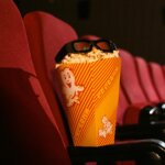 Popcorn box with 3D glasses resting on red theater seats in a dark cinema auditorium.