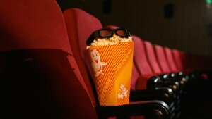 Popcorn box with 3D glasses resting on red theater seats in a dark cinema auditorium.