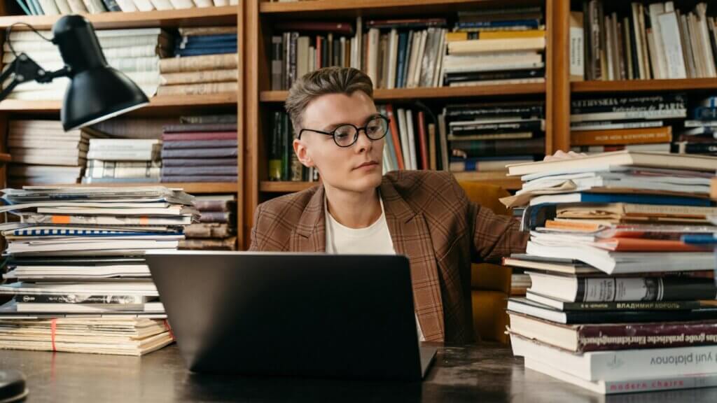 Young man with glasses and plaid blazer working on a laptop surrounded by stacks of books in a library setting