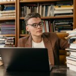 Young man with glasses and plaid blazer working on a laptop surrounded by stacks of books in a library setting