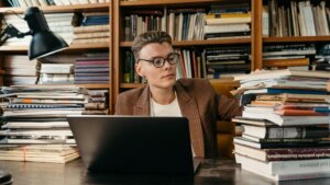 Young man with glasses and plaid blazer working on a laptop surrounded by stacks of books in a library setting
