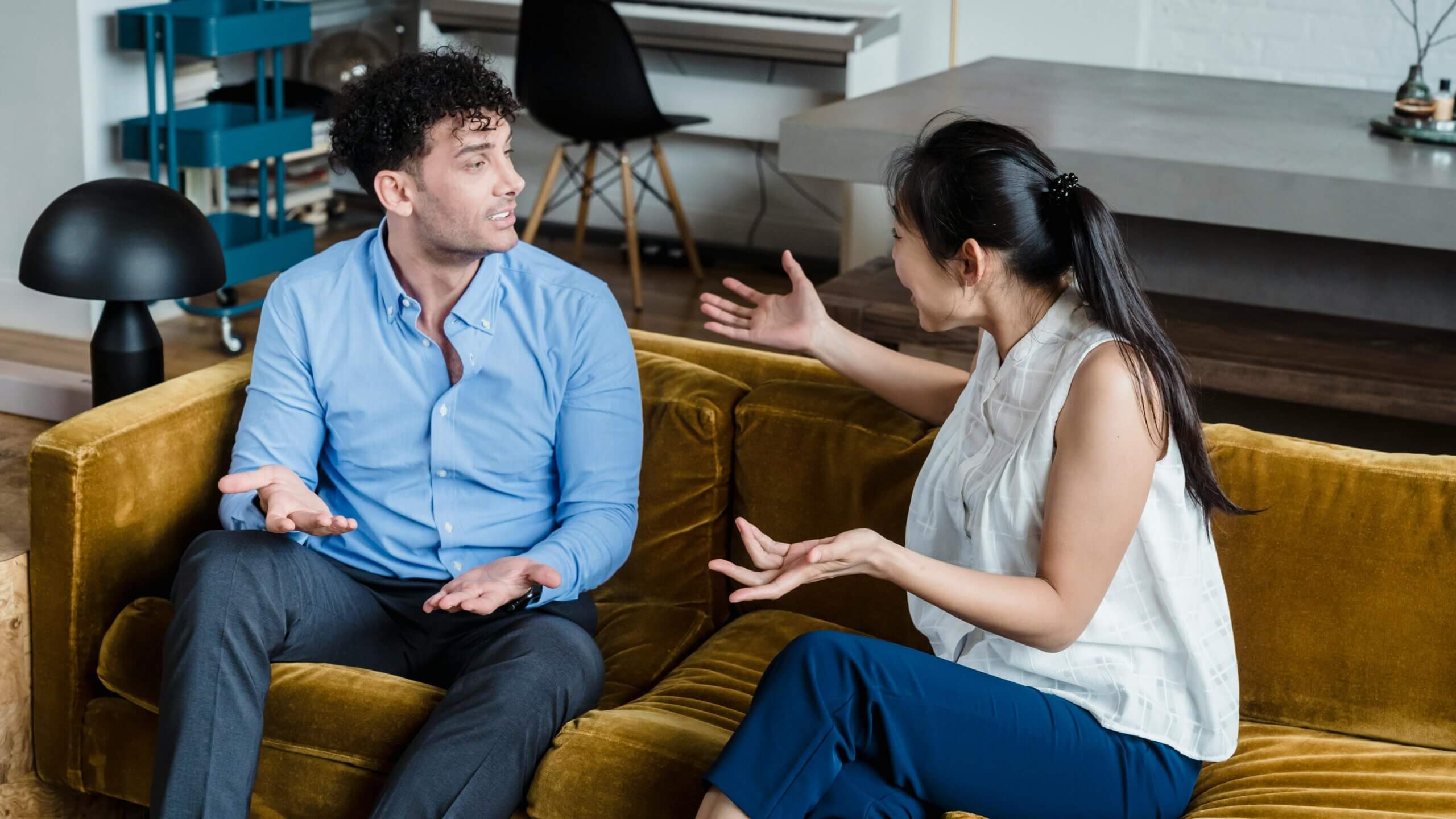 Man in blue shirt and woman in white blouse having a discussion on a mustard yellow couch in a modern living room