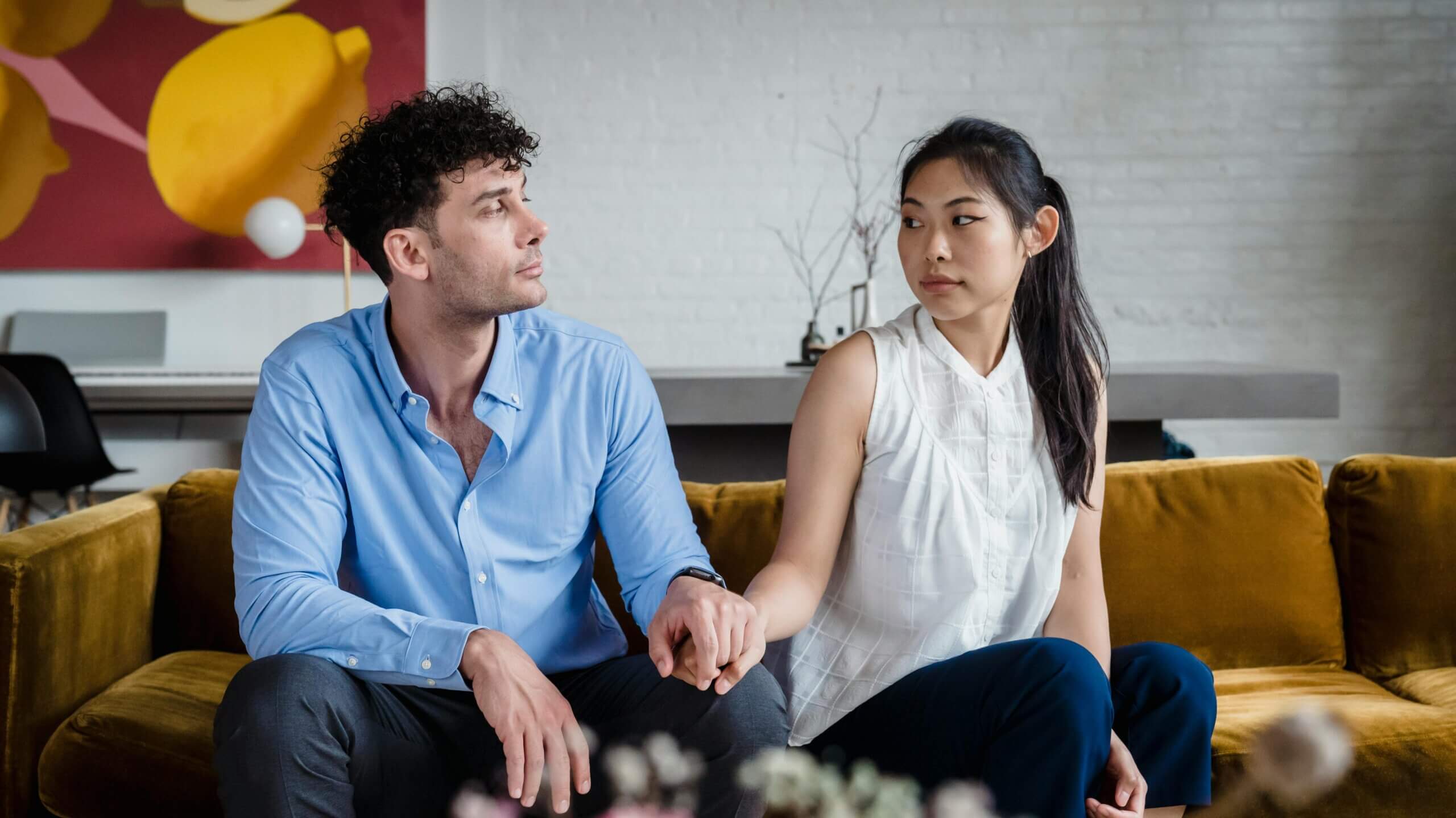 Man and woman sitting on a mustard sofa holding hands and looking away from each other in a modern living room