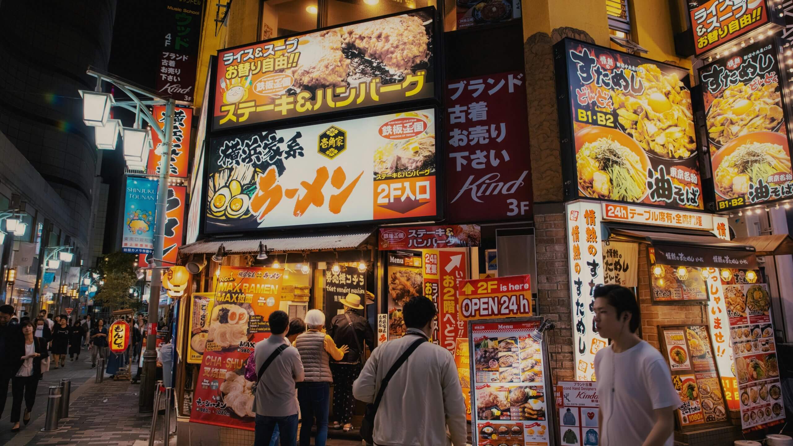 People queue outside illuminated ramen and steak restaurant signs in a busy Japanese street at night.