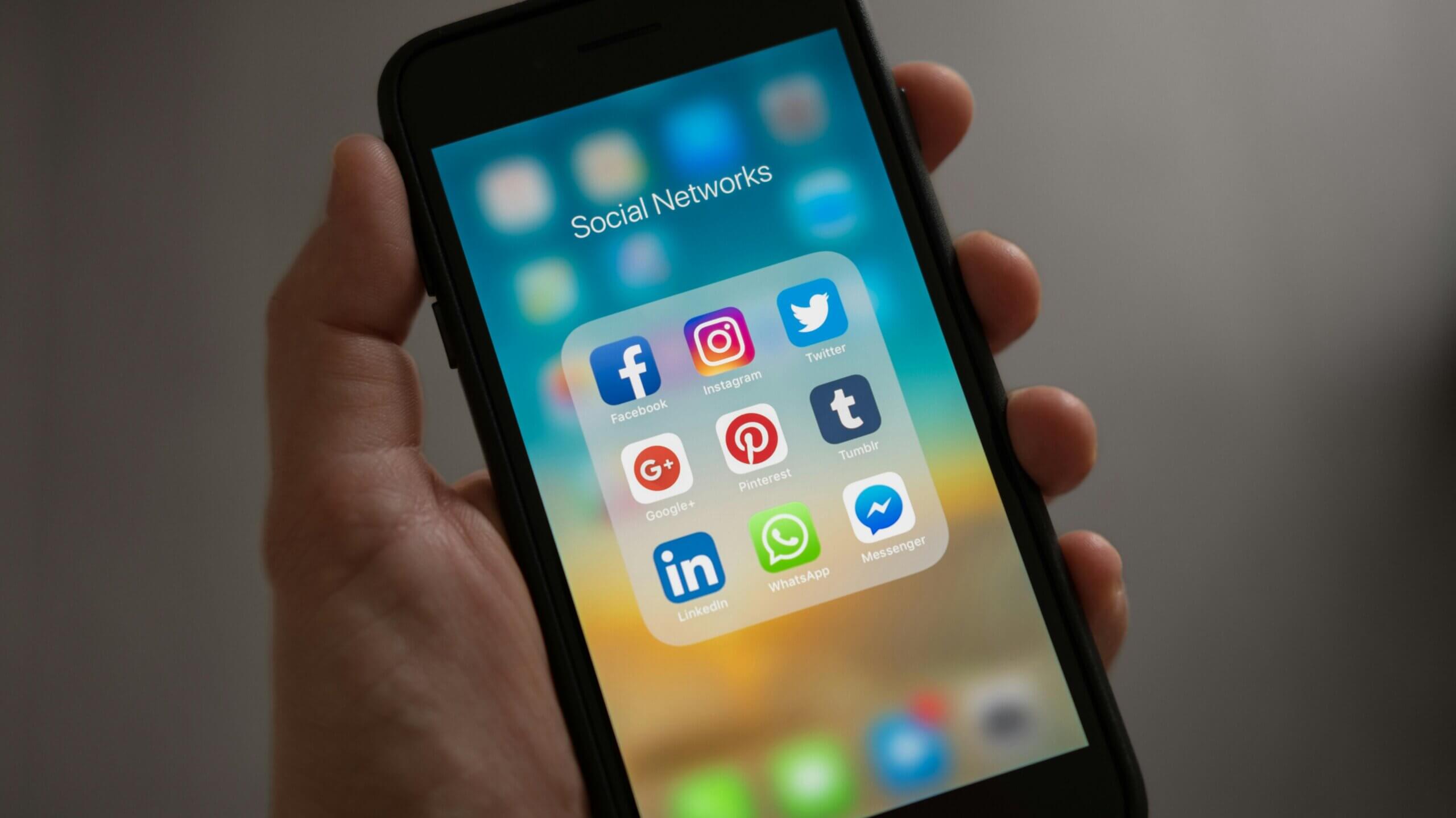 Hand holding smartphone displaying social media apps Facebook, Instagram, Twitter, Google+, Pinterest, Tumblr, LinkedIn, WhatsApp, Messenger.